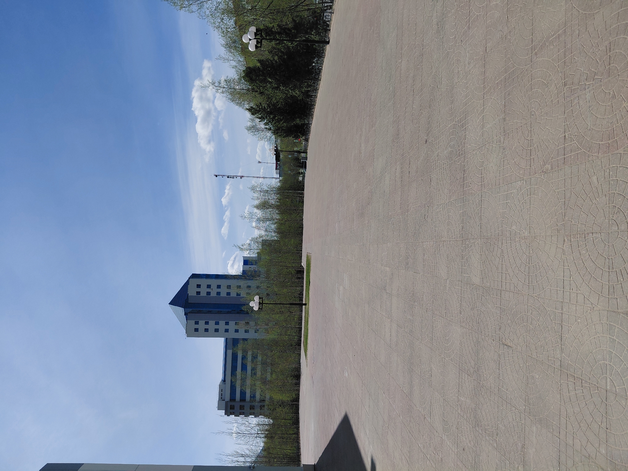 The image displays an outdoor urban landscape, likely a park or public square, under a clear, bright sky. The perspective is slightly elevated, looking across a paved area towards buildings and trees.
The right side of the frame is dominated by a wide, light-colored paved pathway. The paving stones feature an intricate, repetitive pattern of interlocking circles and radial lines. A distinct dark shadow is cast across the bottom left portion of this pathway, indicating bright sunshine and the position of the sun.
To the left of the paved area, a narrow strip of green grass borders a line of trees. Many of these trees appear to be in early spring, with bare branches or just beginning to show light green foliage. Behind this line of trees, several multi-story modern buildings are visible. These buildings are characterized by their light-colored facades, possibly white or light grey, with prominent blue sections, particularly on their roofs and around their windows. The windows are regularly spaced and appear dark, possibly reflective.
Along the edge of the paved path and near the trees, several black streetlights with three spherical lamps are spaced out. In the distance, a tall, slender telecommunication tower with visible antennae can be seen against the sky.
The sky is a vibrant blue with scattered white, fluffy clouds, suggesting clear and sunny weather. There are no people visible, and no specific activities are taking place, creating a calm and static scene. No discernible text is present in the image. The overall impression is one of a tidy, modern public space, set in Nadym, Russia.