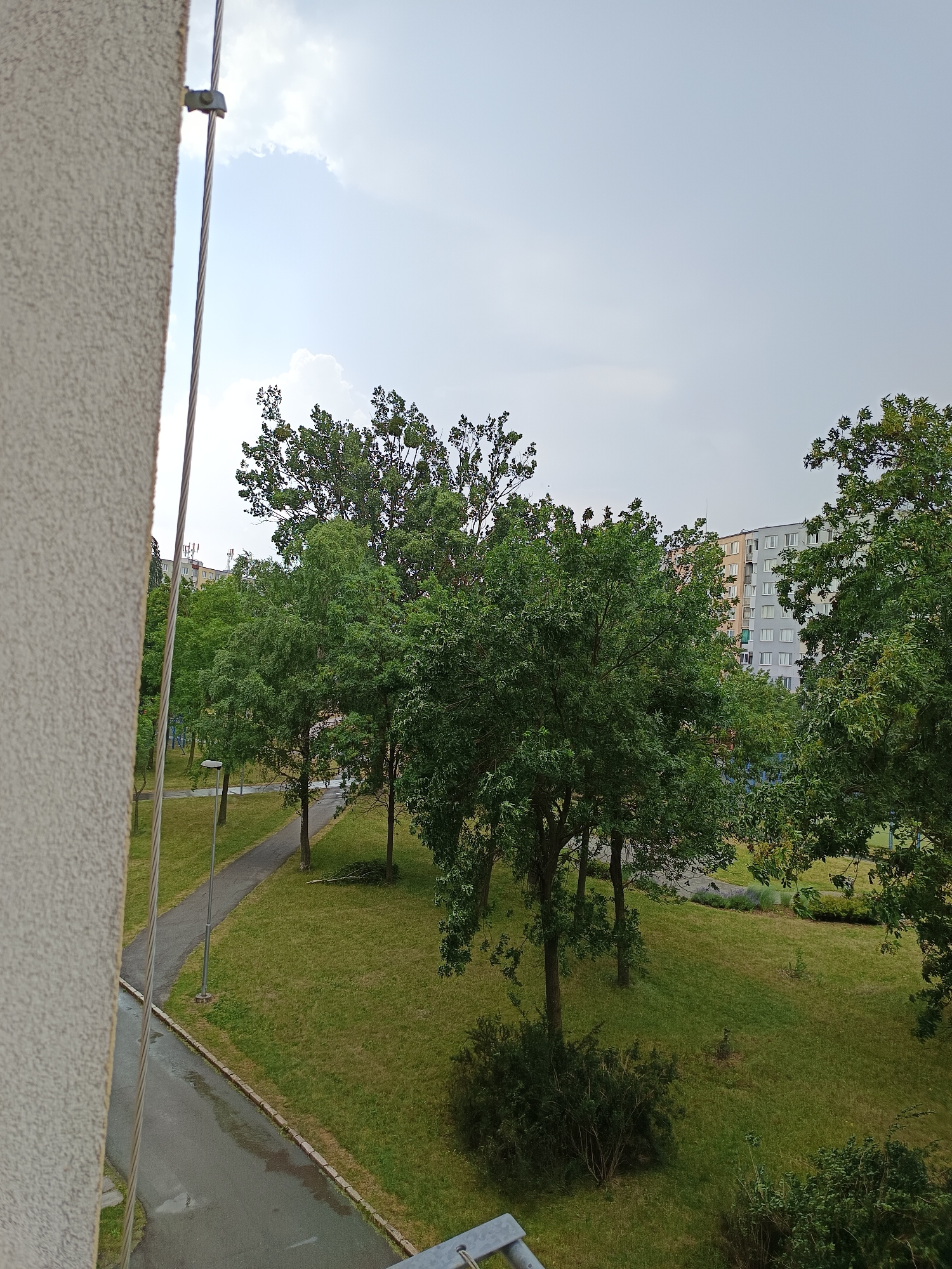 This is a medium-angle, high-definition image taken from a balcony or window, looking down and outwards onto a green park area with apartment buildings in the background. The scene is located in the city of Košice, Slovakia, likely in a residential neighborhood such as Sídlisko III or Sídlisko KVP, characterized by panelák-style apartment blocks and well-maintained public green spaces.
In the foreground, on the left, a textured, light-colored building facade occupies a significant portion of the left edge. A metal cable, likely for electrical or communication purposes, runs vertically along this facade, secured by a metal clamp. The cable extends upwards and to the right.
The central and right portions of the image reveal a park with lush green grass. Several mature trees with dense green foliage are scattered throughout the park, creating a shaded and natural environment. Some smaller bushes and young trees are also visible. A paved pathway, appearing wet and reflective, winds through the park, suggesting recent rain. A lamppost stands along the pathway.
In the background, several multi-story apartment buildings with multiple windows are visible. These buildings are typical of Eastern European urban residential areas. The sky is overcast with light grey clouds, indicating a subdued lighting condition. No people or animals are visible in the scene. The overall impression is of a quiet, urban residential setting with a focus on green spaces.