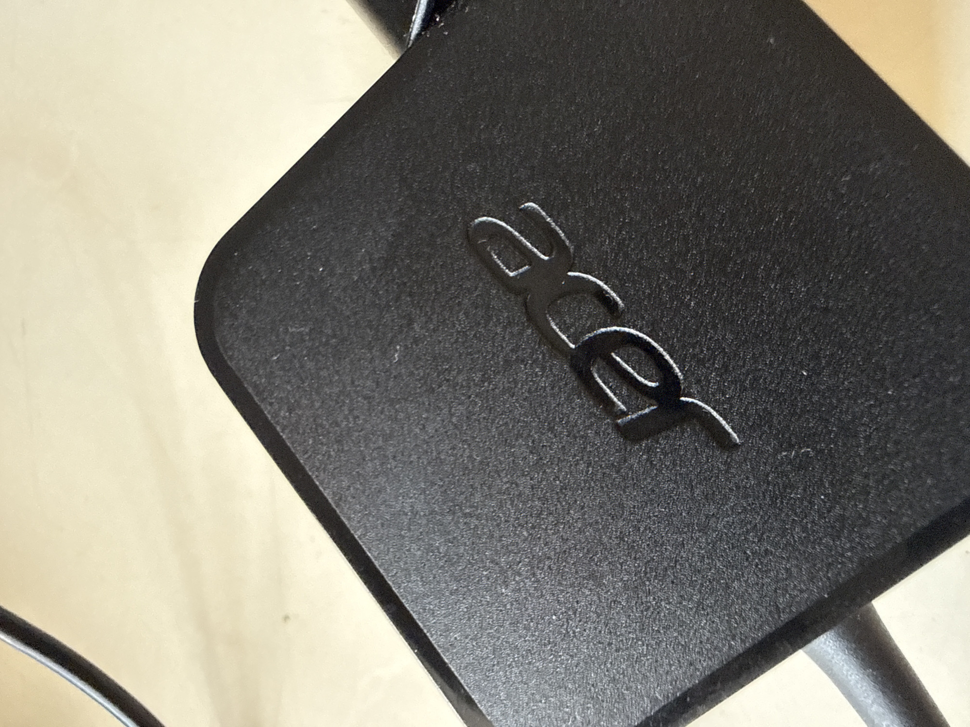 The image shows a close-up of a black Acer power adapter. The Acer logo is embossed on the top surface of the adapter in a stylized font. The adapter has a textured surface and is positioned on a light-colored, possibly wooden, surface. Part of a black cable is visible extending from the top left corner of the adapter. The lighting suggests the image was taken indoors, possibly in a home or office environment.