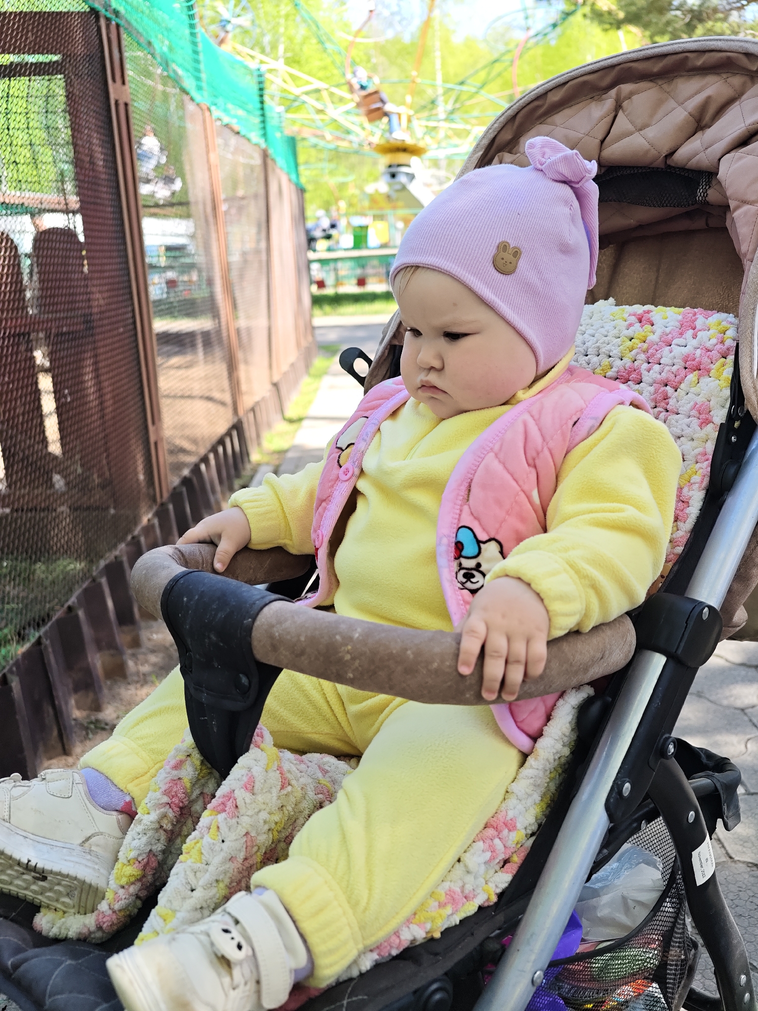 A toddler sits in a stroller on a paved area near a metal fence. The child wears a light pink hat with a small bunny patch, a yellow fleece tracksuit, and a light pink quilted vest. White sneakers with lilac socks peek out from the bottom of the yellow pants. The stroller has a textured blanket and a padded handlebar in front of the child. A mesh bag containing various items is visible beneath the stroller seat. In the background, beyond the metal fence, is a carousel-type amusement ride, indicating a park or amusement area. The location is in the city of Chaikovsky, Russia.