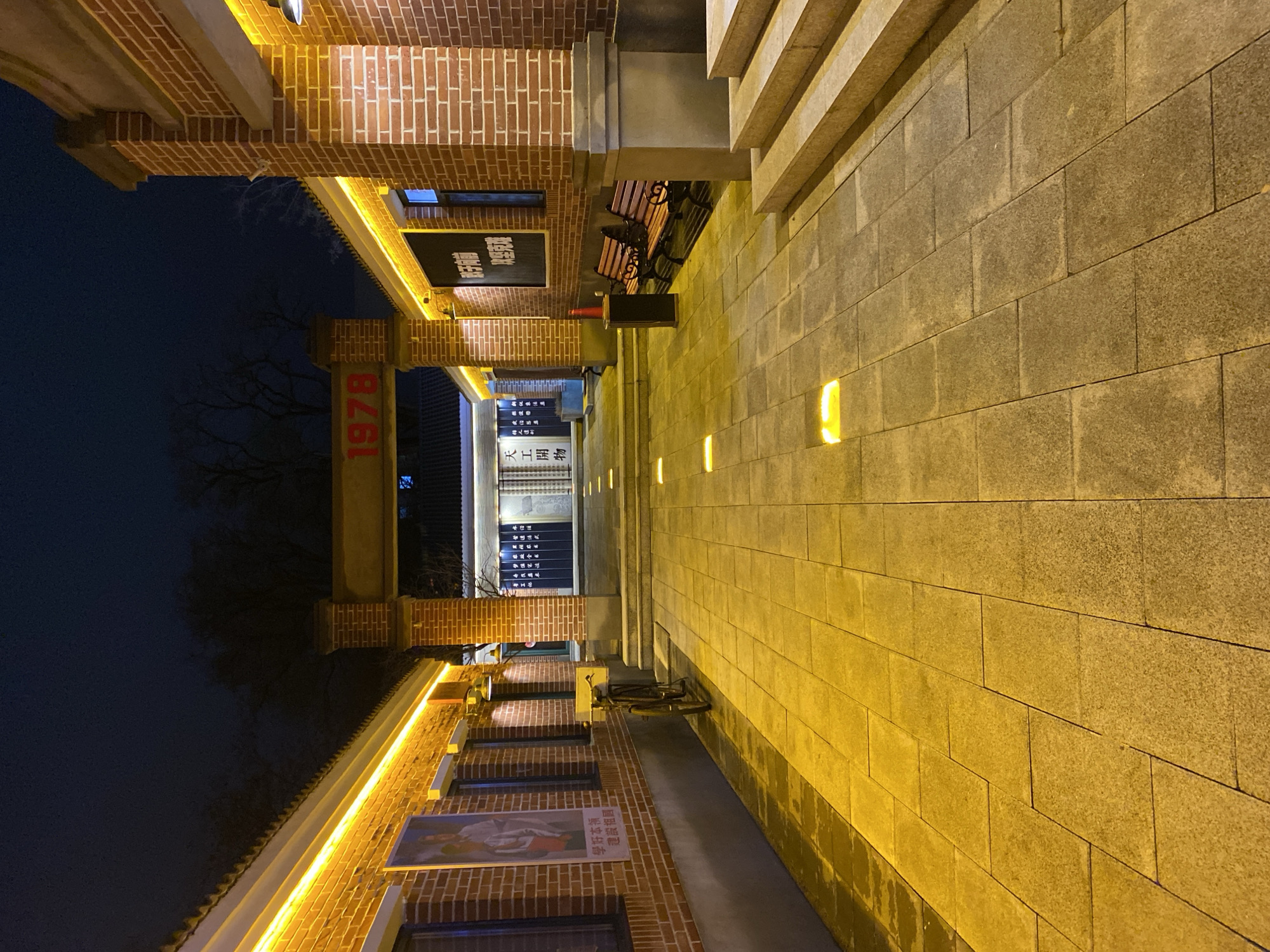 The image depicts an outdoor scene in Zhongtun, China. The foreground shows a wide walkway constructed of light-colored rectangular stones. Low-level lighting fixtures are embedded in the walkway, casting a yellow glow. To the left, a brick building with architectural details including a red number "1978" on a pillar. Signs with Chinese characters are visible on the building. Benches are present along the walkway. The background features trees and a dark sky, suggesting it is either nighttime or a dimly lit area.