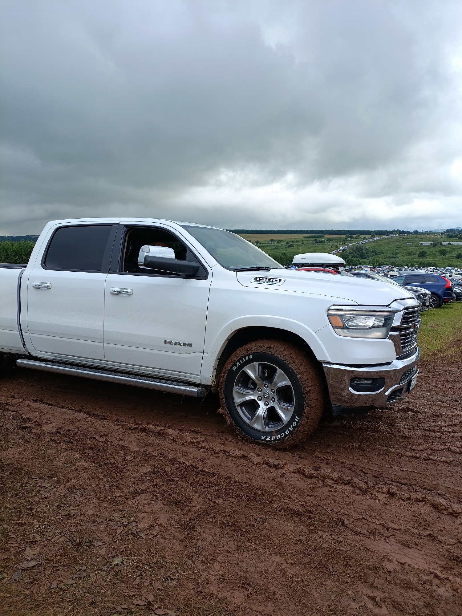 A white Ram 1500 pickup truck is parked on a muddy track, facing right. The truck has a crew cab and is covered in mud, particularly on the front tire and fender. The tire has "ROADCRUZA" visible. Chrome trim is present on the door handles, side mirrors, and front grille. A white cargo box is mounted on the roof of the truck. In the background, a large, crowded parking lot is visible on a grassy hill, with many cars parked in rows. The sky is overcast with heavy, gray clouds. The location is identified as Lagunovo, Russia.