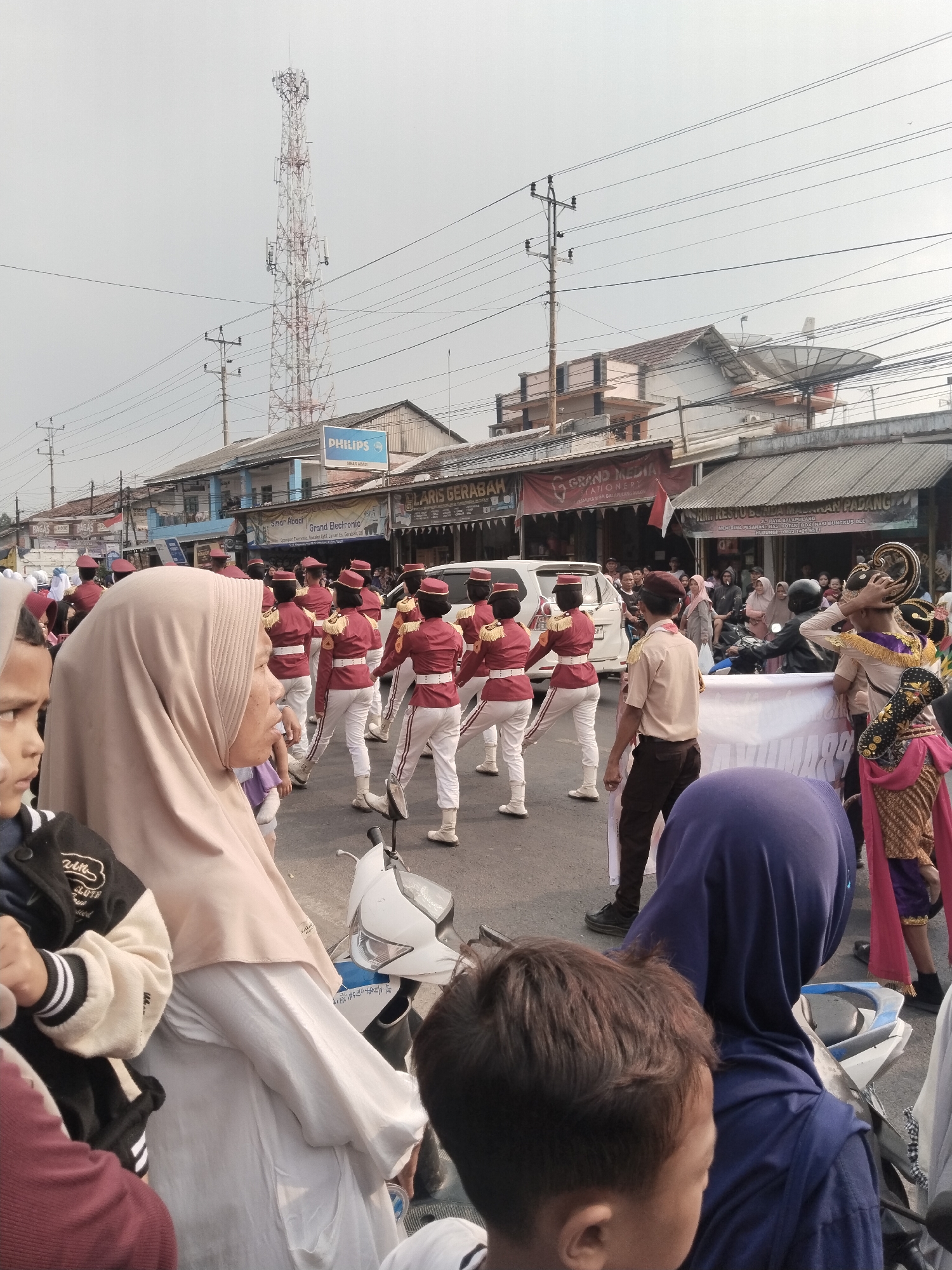The image depicts a street scene in Balapulang Kulon, Indonesia, likely during a parade or cultural event. A marching band, composed of individuals in red jackets with white pants and hats, is the central focus of the procession. Several bystanders, including children and adults, are observing the parade. Some are standing by the roadside, while others are seated on or near motorcycles. Buildings line the street, some of which appear to be shops with visible signage. A prominent cell tower stands in the background, along with utility poles and overhead wires, indicating an urban or semi-urban setting. The sky is overcast, suggesting a cloudy or hazy day. The overall atmosphere appears festive and communal.