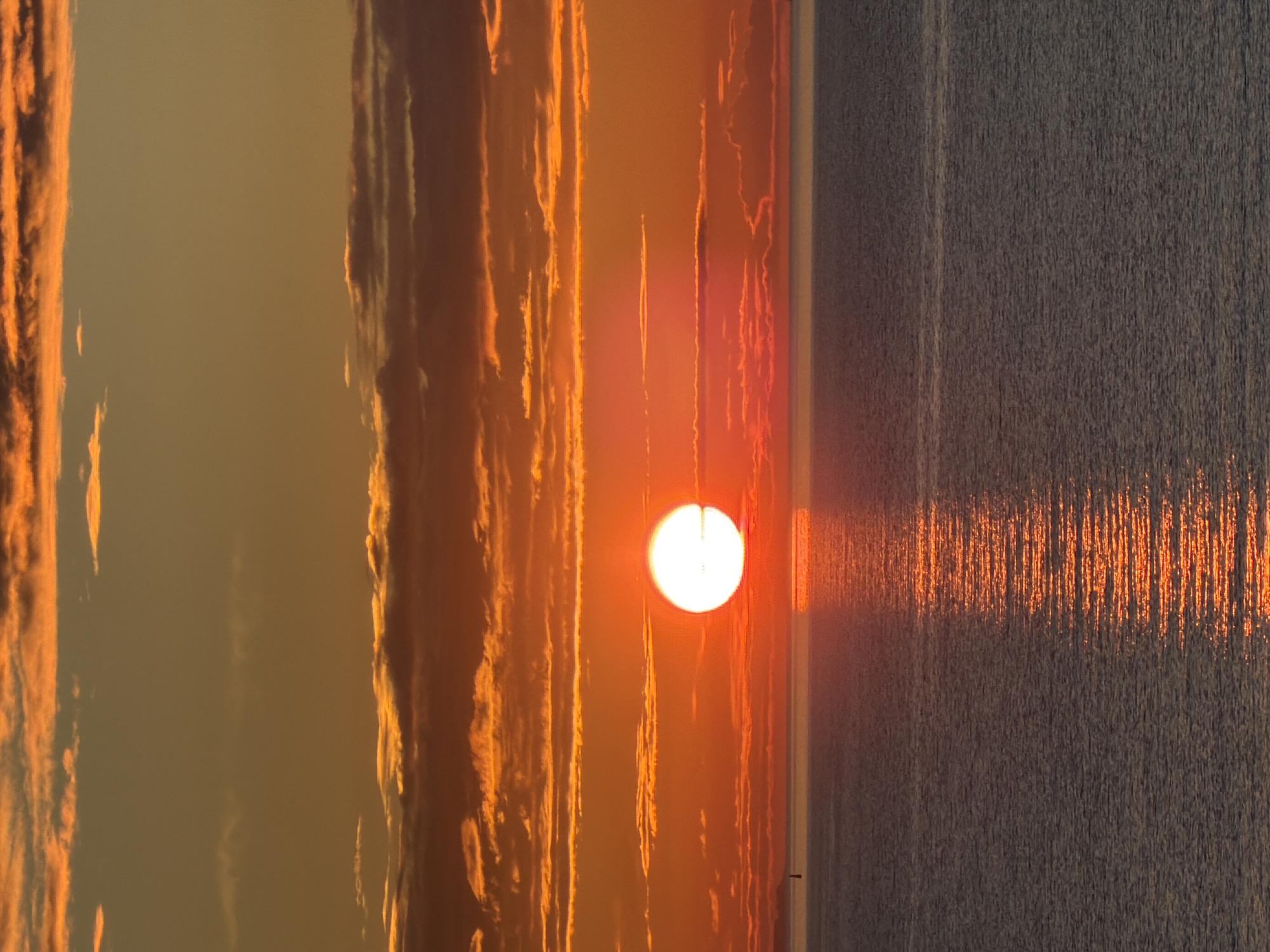 A vibrant sunset over the Baltic Sea is captured from a high vantage point, likely in or near Tallinn, Estonia. The sky is ablaze with hues of orange and yellow, with striated clouds catching the last rays of the sun. The sun itself is a bright, glowing orb partially obscured by a thin layer of clouds, casting a shimmering path of light across the water. The sea is a deep, tranquil blue, reflecting the dramatic colors of the sky. In the distance, a small sailboat is visible on the left, and a faint landmass can be seen on the horizon. The overall impression is one of serene beauty and the grandeur of nature.