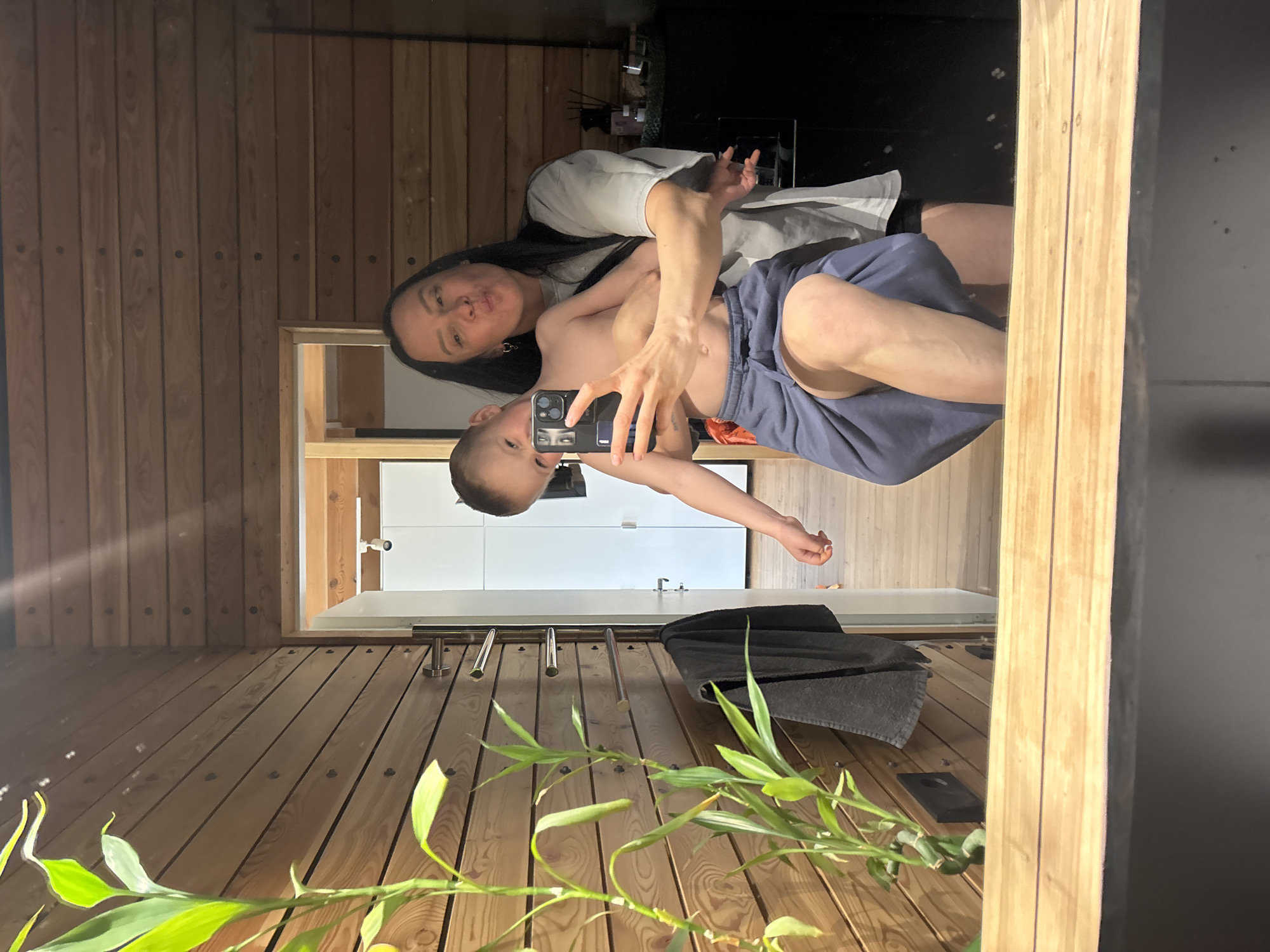 A woman and a child are taking a selfie in a bathroom in what appears to be a home. The woman is wearing a white shirt and has long dark hair. She is holding the child, who is shirtless and wearing blue shorts. The child is holding the phone, seemingly capturing the selfie. The bathroom has wooden walls and flooring. In the reflection, there is a white bathtub and sink. In the foreground, a potted plant is visible. Given the location, the image was likely taken in Chaikovsky, Russia.