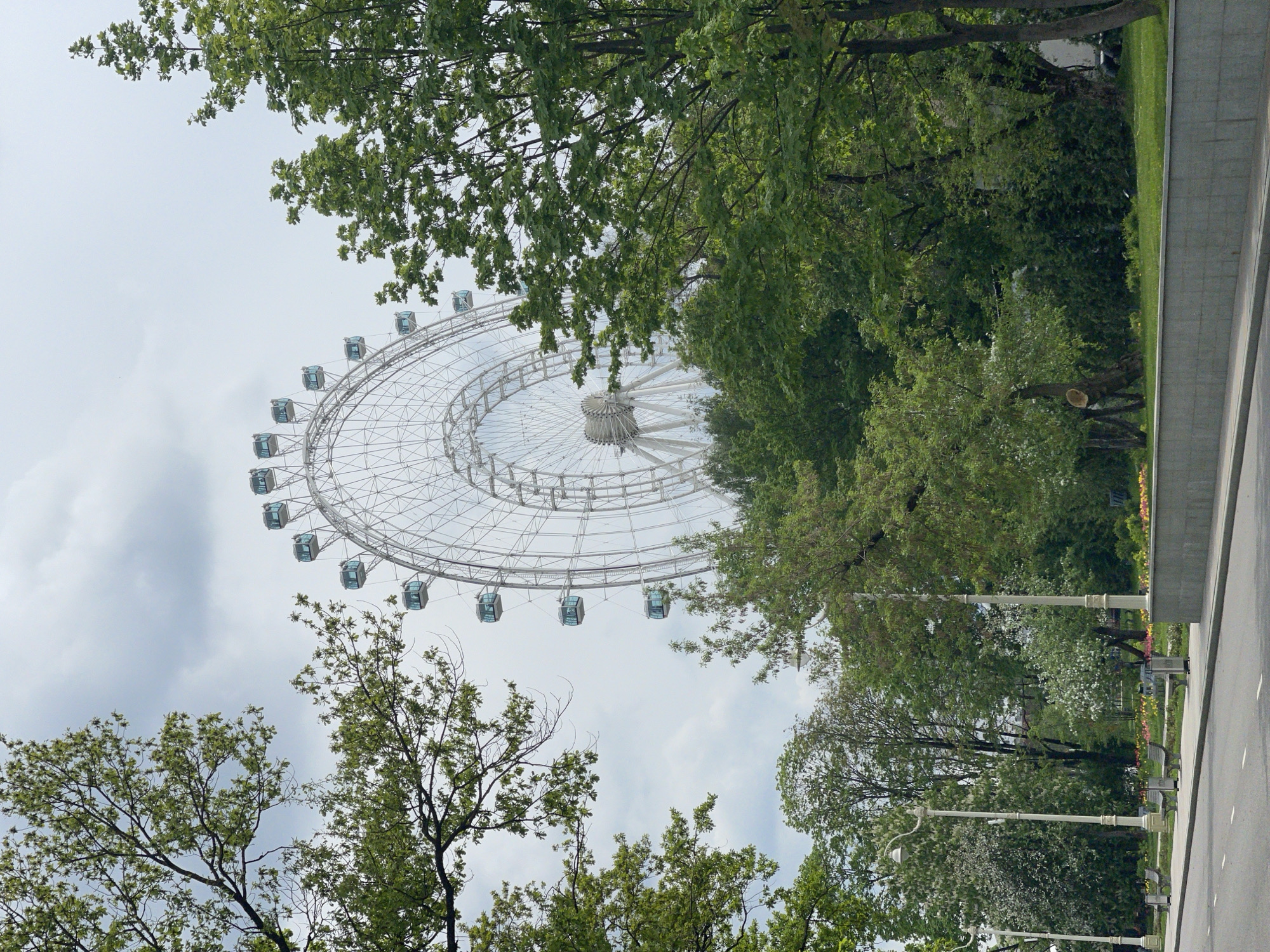 A low-angle view captures a Ferris wheel situated behind trees in a park setting. The Ferris wheel is white, featuring multiple passenger cars and a central hub. Trees with green foliage are positioned in the foreground and midground, partially obscuring the Ferris wheel. The sky is overcast. A road runs along the right edge of the scene, beside trees, lamps, benches, grass, and a wall. The location is Unknown, Unknown.