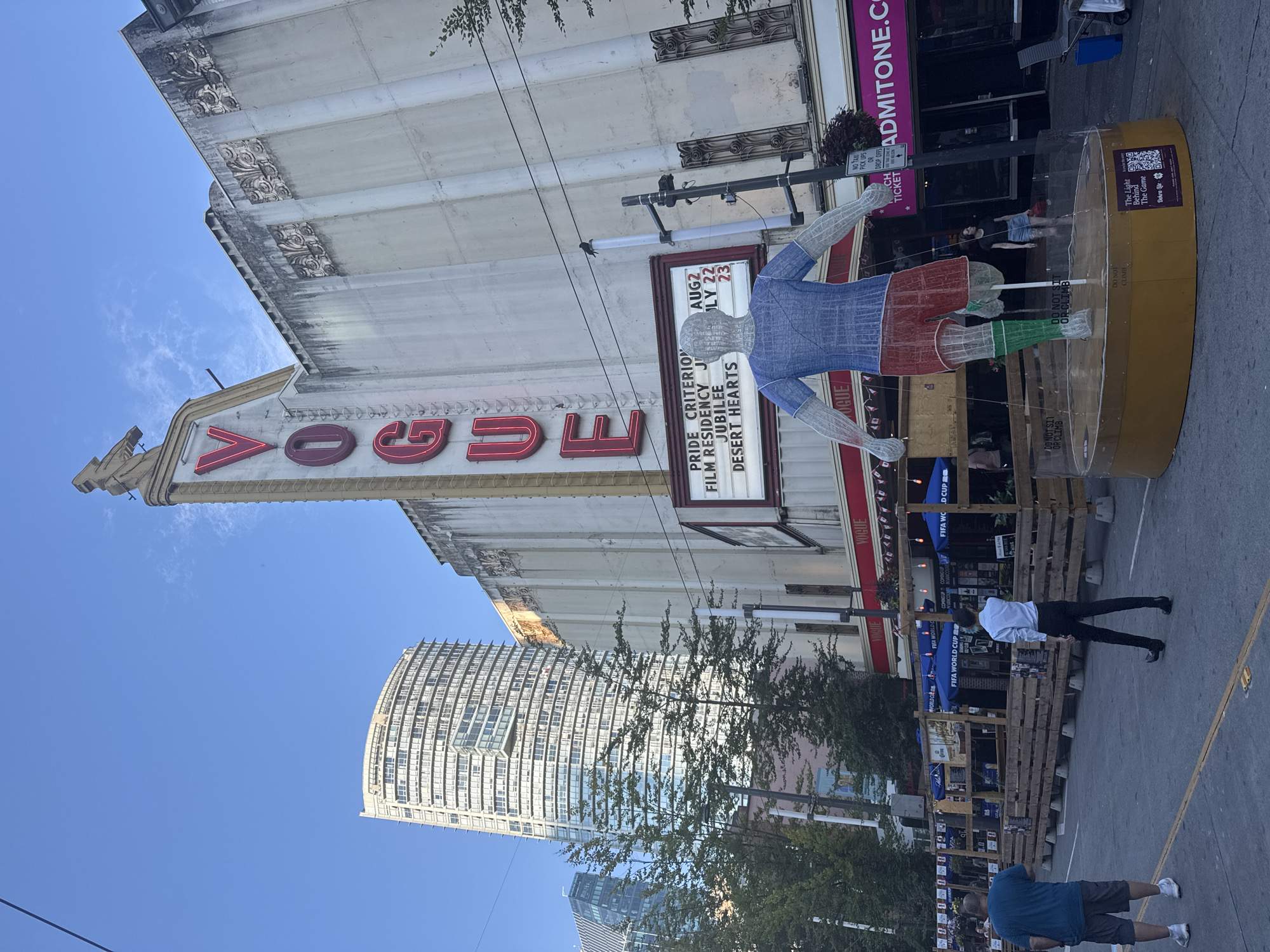The image depicts the Vogue Theatre in Vancouver, Canada, on a clear day. A large wireframe sculpture of a person wearing a blue shirt and red shorts with green socks stands in the foreground, positioned on a circular, yellow platform. The sculpture is surrounded by a transparent barrier. In the background, the iconic "VOGUE" sign in red neon lettering is prominently displayed on the facade of the theater. Below the sign, a marquee lists movie titles, including "PRIDE CRITERION," "FILM RESIDENCY," "JUBILEE," and "DESERT HEARTS," with dates "AUG 2," "JULY 22," and "23." The theater's facade is light-colored with decorative architectural elements. To the left of the theater, a modern, tall, curved skyscraper with numerous windows rises into the sky, characteristic of Vancouver's downtown architecture. The street in front of the theater features outdoor seating arrangements with wooden barriers and blue umbrellas, suggesting a dining or event space. Two men are walking away from the camera on the sidewalk, one wearing a blue t-shirt and shorts, the other a white shirt and black pants. Other individuals are visible further in the background near the theater entrance. The overall scene conveys a vibrant urban environment, likely in the entertainment district of Vancouver.