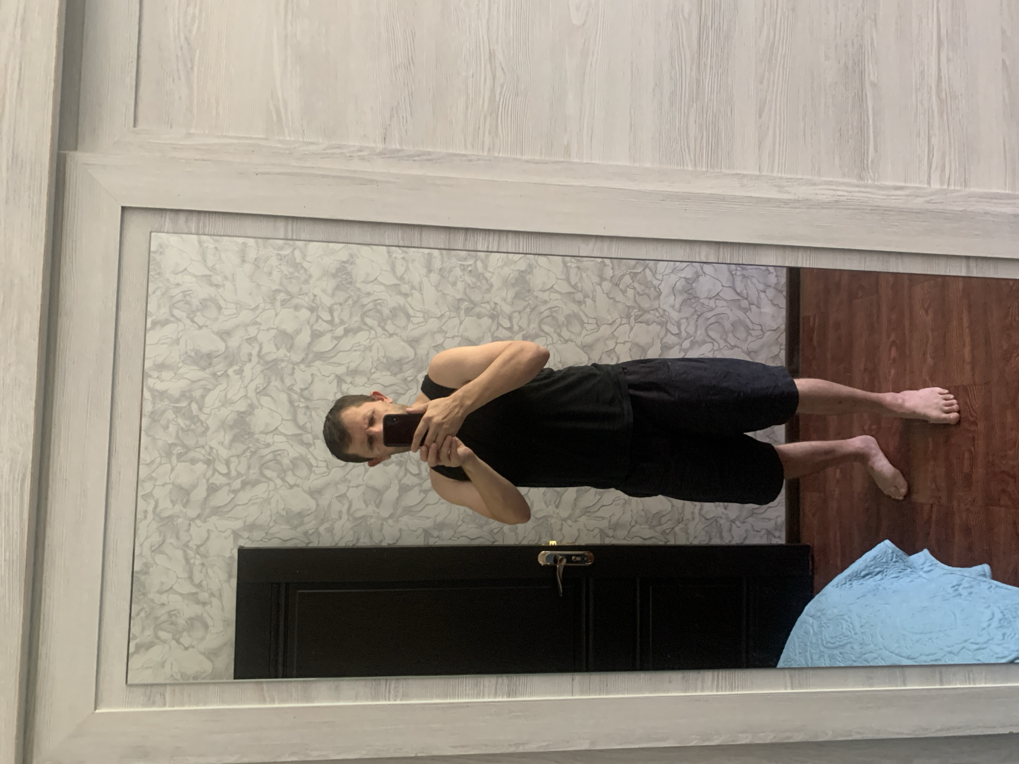 A young man is taking a mirror selfie in a bedroom. He is wearing a black tank top and black shorts. He is barefoot. In the reflection, a black door with a silver handle is visible on the left side of the frame, and a blue quilted blanket is partially visible on a bed on the far left. The room has light-colored, wood-grain patterned walls and dark wood flooring. The mirror is framed by a light-colored, textured border. The setting is consistent with a residential interior in Tashkent, Uzbekistan.