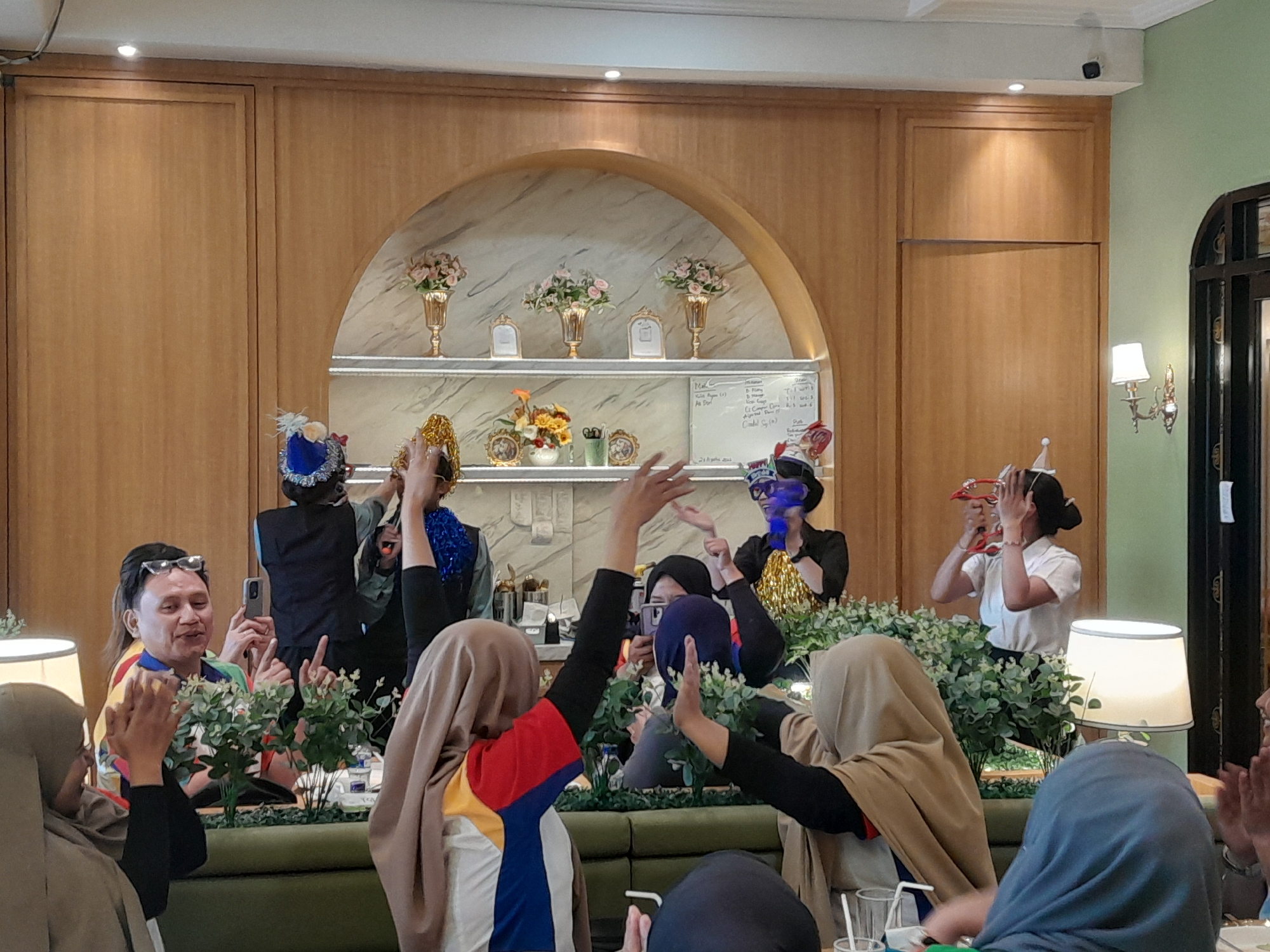 In a brightly lit indoor space, possibly a cafe or restaurant with wood-paneled walls and a decorative arched shelf displaying vases with flowers and framed pictures, a group of people are gathered around a long, green booth. Many are wearing festive party hats and headwear. In the foreground, a person wearing a beige hijab is clapping, and another in a blue hijab is visible from the chest up. To the left, a man wearing glasses and a colorful striped shirt is holding up a smartphone, appearing to take a picture or video. Behind him, several individuals are engaged in activities, with some raising their hands. One person on the right is holding what appears to be a tambourine. The overall atmosphere suggests a celebratory gathering, possibly a birthday or other special occasion. The location is identified as Sempusari, Indonesia.