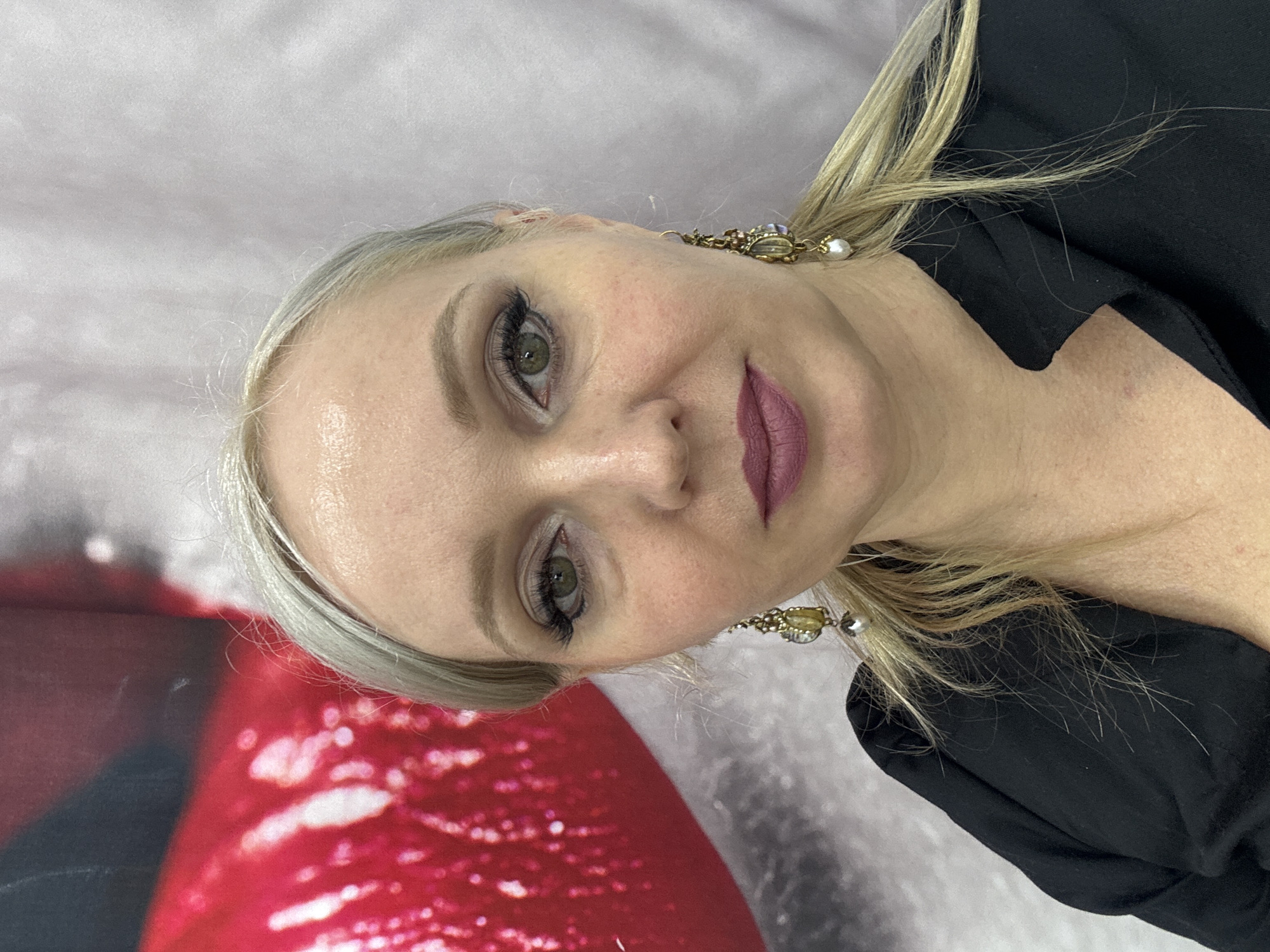 A close-up portrait shows a woman with blonde hair, green eyes, and makeup, including eyeliner and lipstick. She is wearing a black top and ornate earrings. The background consists of a blurred image featuring a large red shape, possibly lips, and a soft, textured grey surface. The image was taken in Komsomolsk-on-Amur, Russia.
