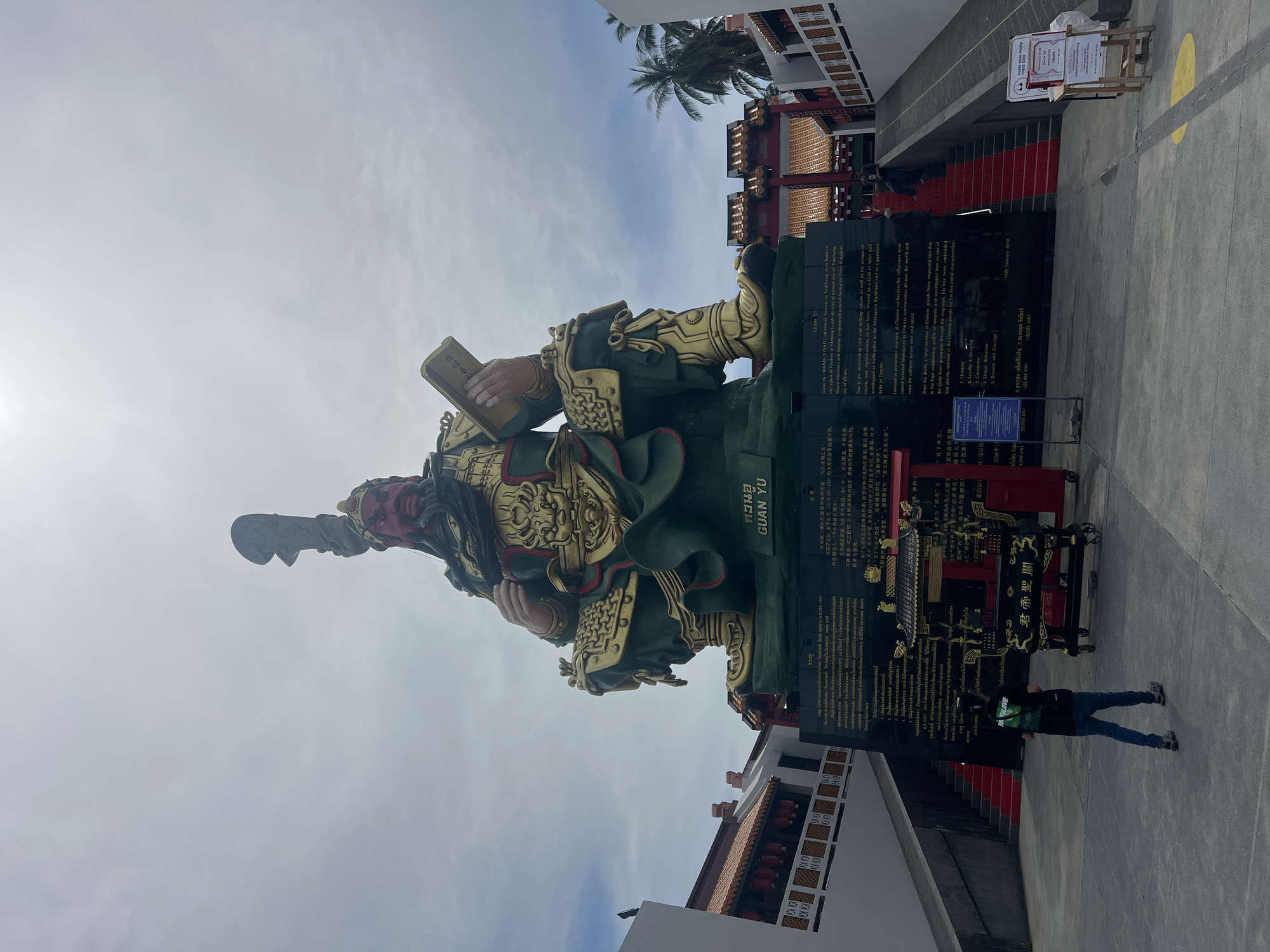 An extremely large, green and gold statue of Guan Yu, a Chinese general and deity, sits majestically under a cloudy sky in Baan Hua Thanon, Thailand. The statue is seated and prominently features intricate gold detailing on its armor and facial features, with a large weapon held aloft and a book in its hand. At the base of the statue is a large black plaque with Chinese and Thai inscriptions, likely providing historical or religious information. In front of the plaque, a person wearing a black shirt and blue jeans walks away from the camera towards the statue, their back to the viewer. To the right and left of the statue, red-carpeted stairs lead up to traditional East Asian-style buildings with red roofs and decorative elements. Palm trees are visible in the background, suggesting a tropical location. A small wooden stand with signs is positioned on the right side of the frame, near the bottom of the stairs. The overall scene depicts a significant cultural and religious landmark.