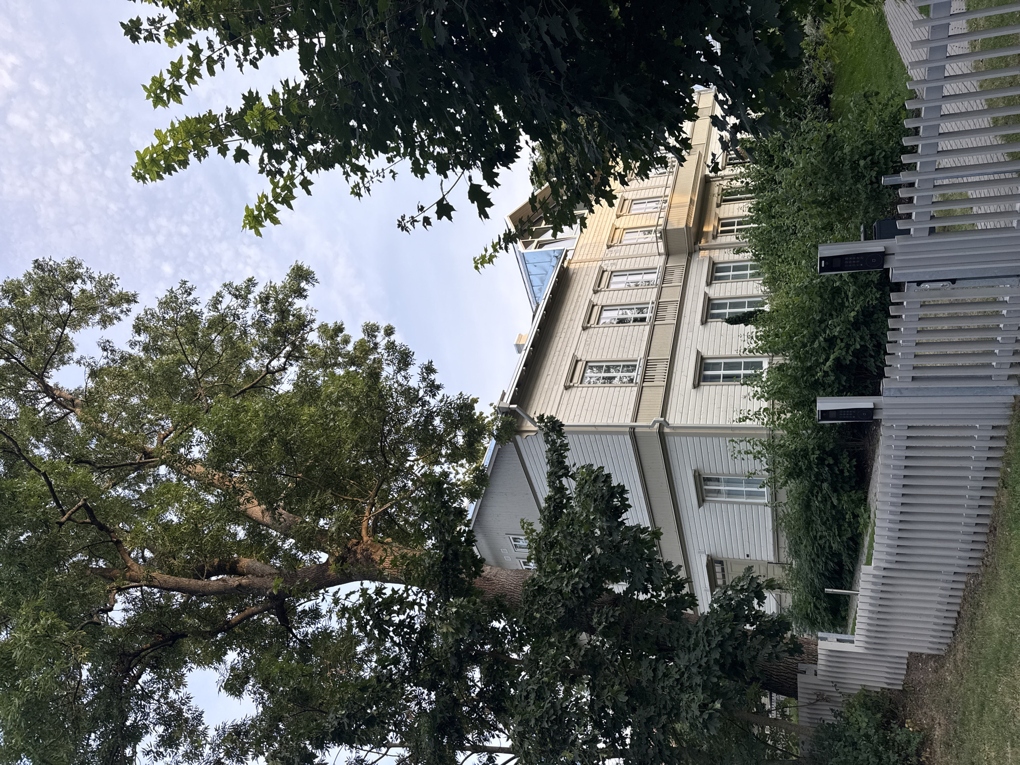 The image shows a light-colored, two-story wooden house with a balcony on the upper floor. The house is surrounded by lush green trees and bushes. A light-colored wooden fence runs along the front of the property, separating it from a paved walkway and a grassy area. The sky is partly cloudy, with a mix of blue sky and white clouds. The house appears to be in a residential area, possibly in the city of Tallinn, Estonia, given the architectural style and the surrounding vegetation. There are no people visible in the image. The lighting suggests it is either early morning or late afternoon.