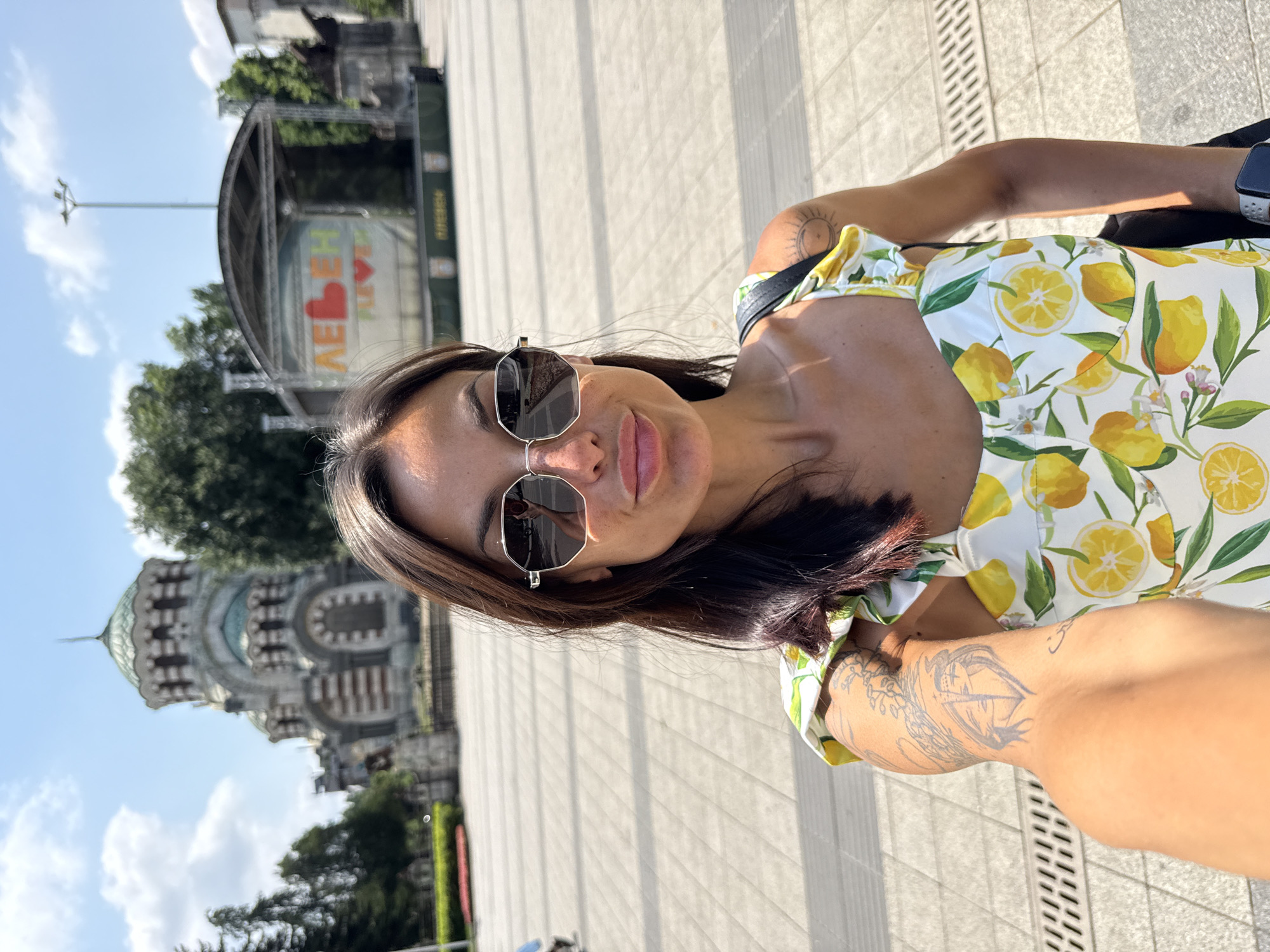 A woman is taking a selfie outdoors in front of a large, ornate building with a green dome. She is wearing a white dress with a lemon print, sunglasses, and has tattoos on her arms. Behind her, a stage with a banner reading "PLEVEN LOVE" is visible. The setting appears to be a public square in the city of Pleven, Bulgaria.