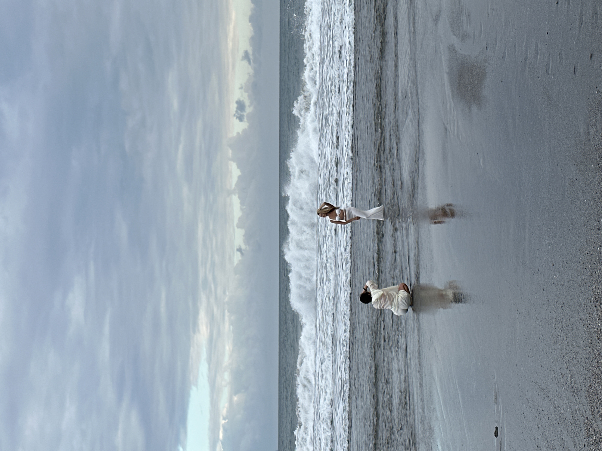 In North Kuta, Indonesia, on a beach under a cloudy sky, two individuals are engaged in a photoshoot. The scene is set at the water's edge, with gentle waves lapping at the shore and larger waves breaking further out. The sand appears wet and reflective, mirroring the figures.
One person, positioned to the left and slightly closer to the viewer, is kneeling in the shallow water. They are wearing a light-colored, loose-fitting outfit, possibly a robe or cover-up over shorts, and are holding their hands up to their head. This individual appears to be the photographer, as they are looking towards the other person and holding what could be a camera or phone.
To the right and further into the water stands another person, the subject of the photoshoot. This individual is wearing a white, two-piece outfit, consisting of a halter top and a long skirt. They are posing with one hand touching their head, looking away from the camera. Their stance is elegant and poised.
The ocean is a prominent feature, with its foamy white crests contrasting against the darker blue water. The sky is overcast with a mix of gray and white clouds, suggesting a diffused light condition. The overall atmosphere is serene and artistic, with the natural beauty of the beach serving as a backdrop for the human interaction.