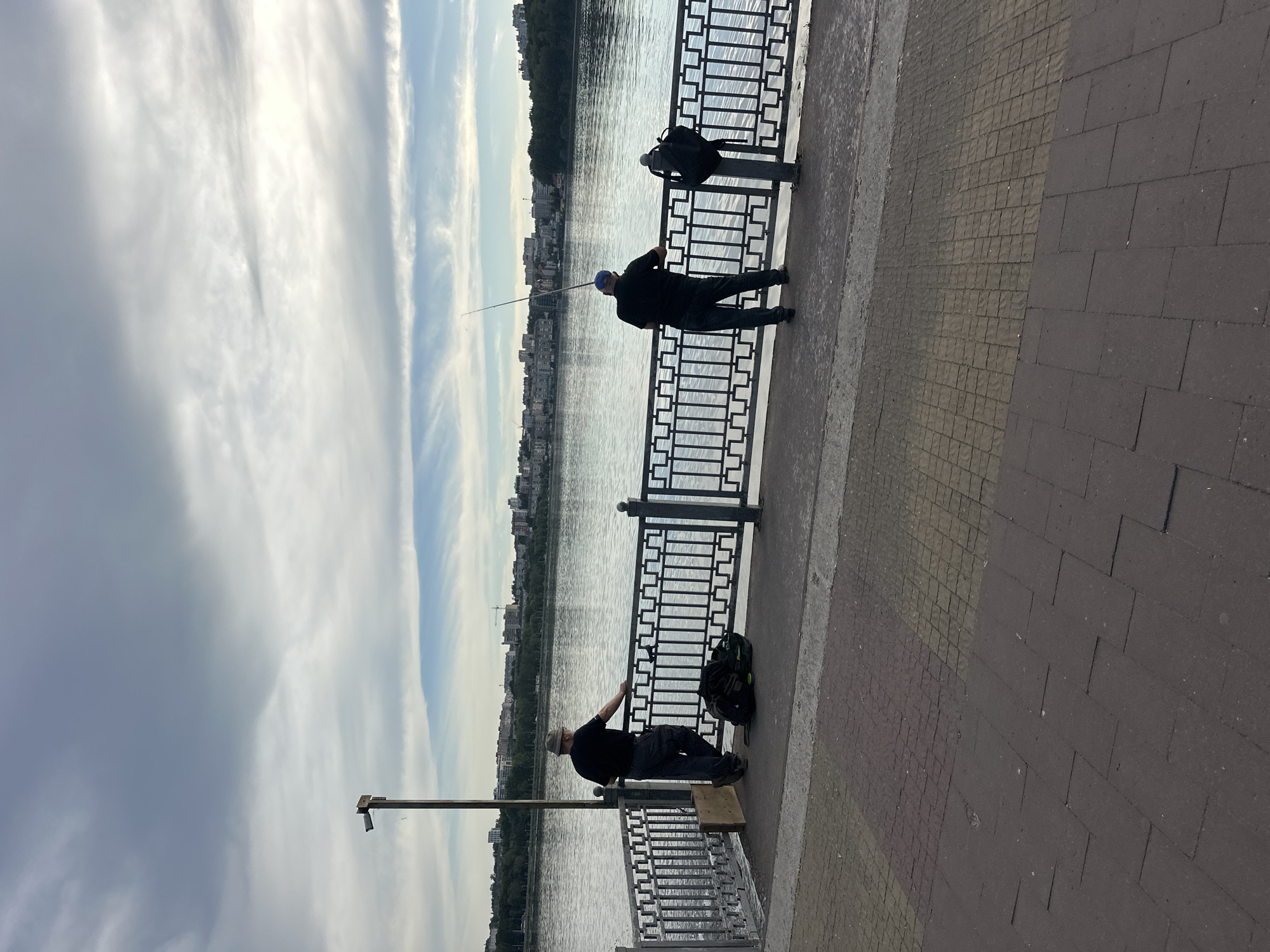 The image depicts a waterfront scene with two individuals fishing from a promenade. In the foreground, a paved walkway features two distinct patterns of paving stones. Beyond the walkway is a metal railing bordering a body of water, likely Ternopil Pond in Ternopil, Ukraine, given the distinctive skyline of buildings and trees on the opposite bank.
Two men are visible on the promenade, both facing away from the camera and towards the water, engaged in fishing. The man on the left, wearing a dark t-shirt and grey pants, is leaning on the railing. Next to him are a dark backpack and a piece of plywood. The man on the right, dressed in a black t-shirt, dark pants, and a blue baseball cap, holds a fishing rod extended over the water. A black backpack is also present near him, attached to a railing post.
The sky above is partially cloudy, with dramatic cloud formations suggesting either an approaching storm or the aftermath of one. The water is calm, reflecting the sky and the distant cityscape. The overall atmosphere suggests a quiet, recreational activity taking place in an urban park setting.