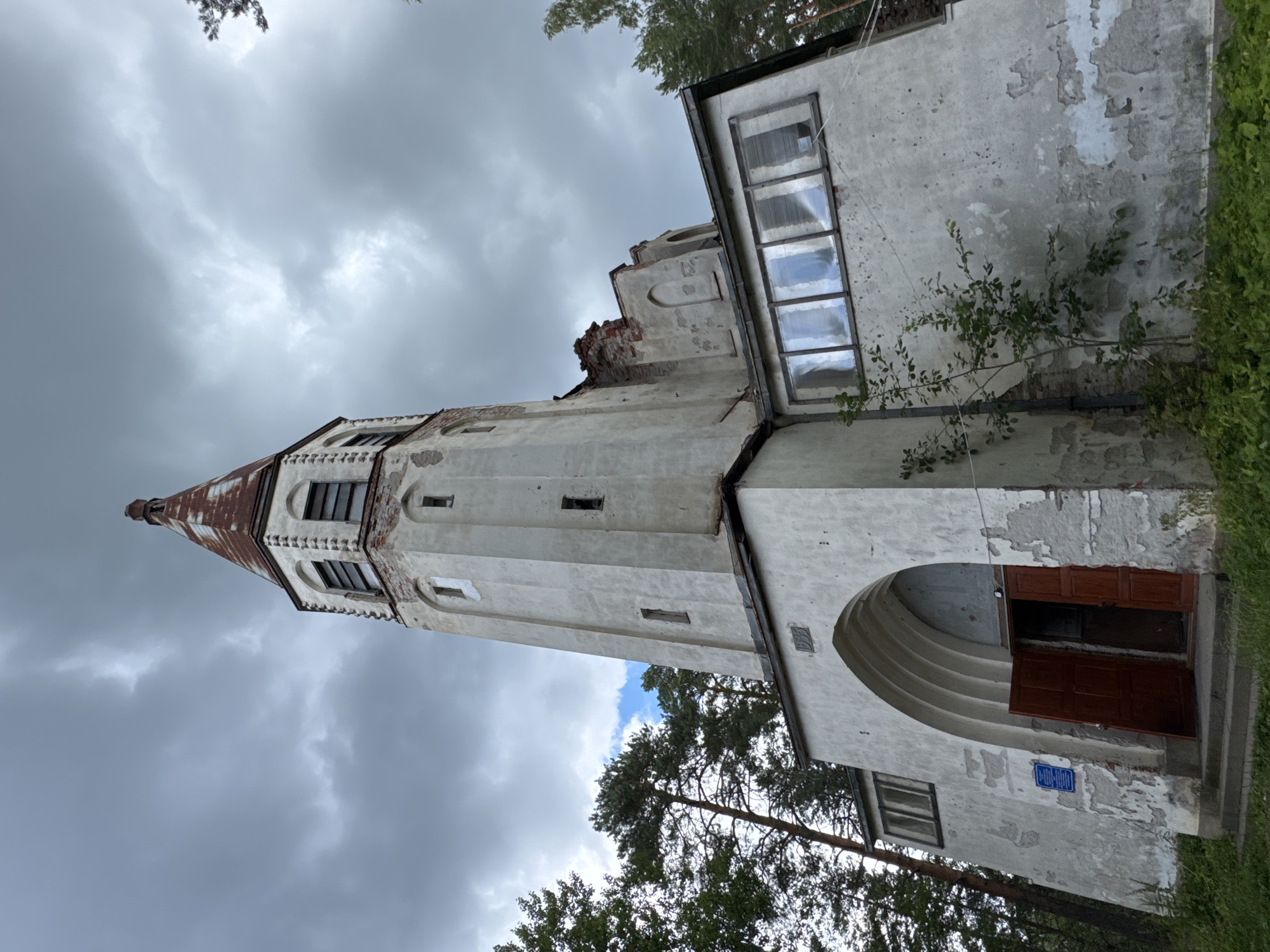 The image shows a low-angle, wide shot of a dilapidated, white, gothic-style tower in Lumivaara, Russia. The tower has a pointed, rusty metal roof and several rectangular windows. The lower section of the tower has a large arched entrance with open wooden doors. The building's stucco exterior is peeling, revealing brick underneath. A small tree grows in front of the building. The sky is overcast with dark clouds.