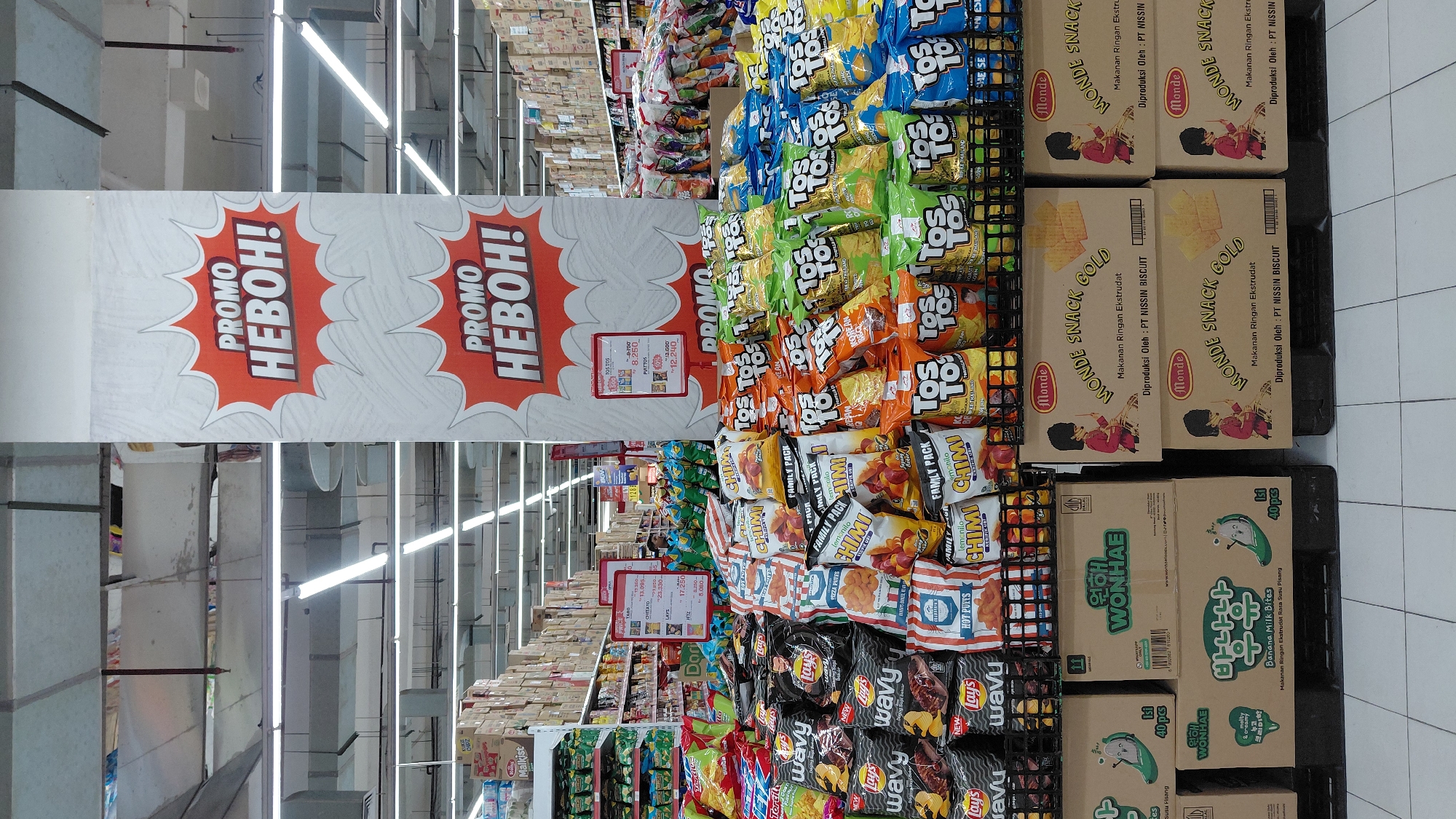 The image shows an aisle in a supermarket, likely in Purwokerto, Indonesia, featuring a prominent display of snack foods. A large vertical banner with the text "PROMO HEBOH!" (meaning "Sensational Promo!") in red lettering on a white background with orange bursts is prominently displayed in the center. Below this, a similar banner is visible. The shelves are stocked with a wide variety of snack bags, including Lay's, Wavy, Chimi, and Tos Tostos chips in various flavors like original, chili, and cheese. Some bags are marked "FAMILY PACK." In the foreground, stacked boxes of "Monde Snack Gold" and what appears to be "Banana Milk Bites" are visible, with smaller text indicating production by PT Nissin Biscuit. The environment is brightly lit by fluorescent lights on the ceiling, and the floor is tiled. The overall impression is a busy retail environment focused on promoting snack products. There are no people clearly visible in the foreground or mid-ground, but a few figures in uniforms can be seen further down the aisle in the background.