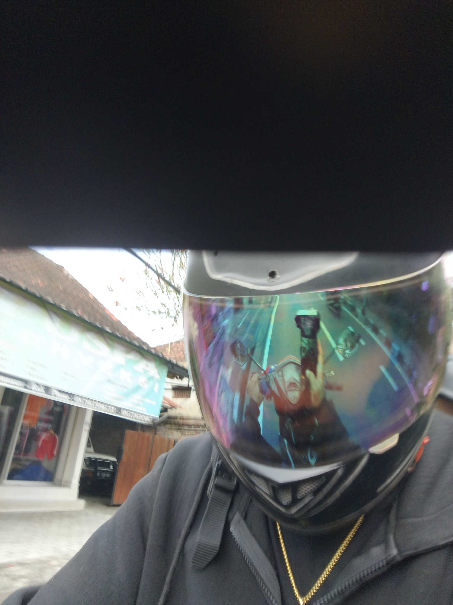 The image captures a first-person perspective of a person wearing a black hooded sweatshirt and a full-face motorcycle helmet with a iridescent visor. The visor reflects a street scene, showing a red motorcycle, a person on the motorcycle holding a phone, and other vehicles and buildings. In the background of the actual scene, a building with a brown tiled roof and a light blue sign is visible. There are also some trees with sparse foliage. The overall impression is that the photo was taken while riding a motorcycle in Denpasar, Indonesia, on a street with shops and residential buildings. The image is slightly blurry, suggesting movement.