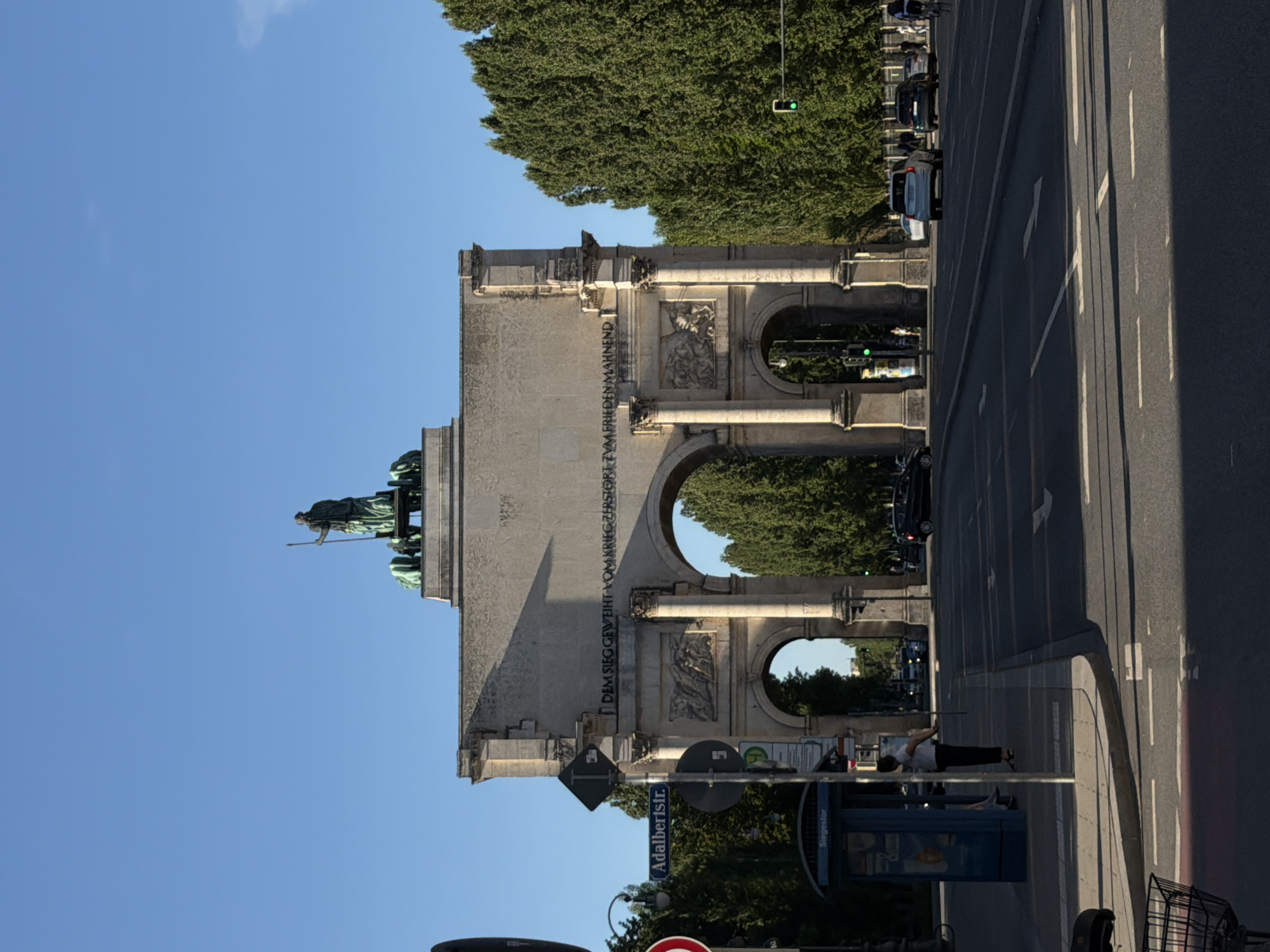 The image shows the Siegestor (Victory Gate) in Munich, Germany, on a clear day. The stone archway is adorned with sculptures and an inscription. A statue of a woman with a spear stands atop the monument. In the foreground, a street with traffic lanes and pedestrian crossings is visible. Cars are driving on the street, and a traffic light shows green. A blue bus stop shelter with the word "Siegestor" on it is located on the sidewalk. A woman in a white shirt and dark pants is standing near the bus stop, holding a long stick. Trees line the street on both sides, and a bright blue sky fills the upper portion of the image.