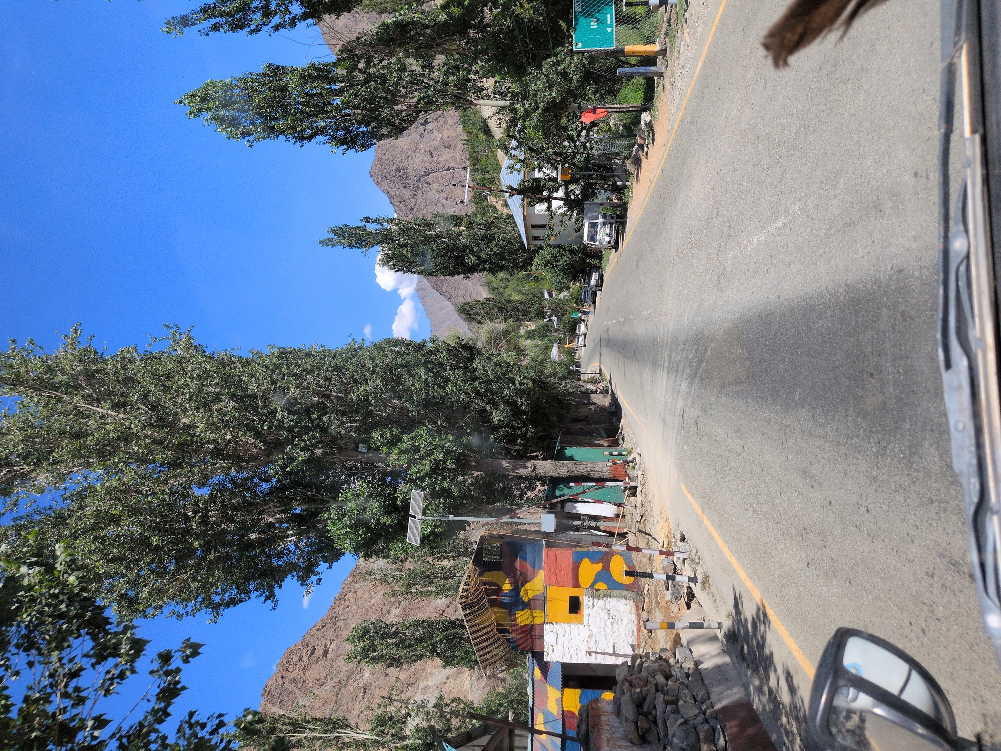 The image shows a view from inside a vehicle traveling on a paved road in a village, likely in Chanigund, India, as suggested. The road has a yellow dividing line. On the left side of the road, there is a building with brightly colored murals, and beyond that, a stone wall and trees. A tall solar-powered lamp is positioned near the building. On the right side of the road, there are more trees, houses, parked cars, and a green road sign. The surrounding environment features dry, rocky mountains and a clear blue sky. The presence of trees and a building suggest a rural or semi-urban setting.
