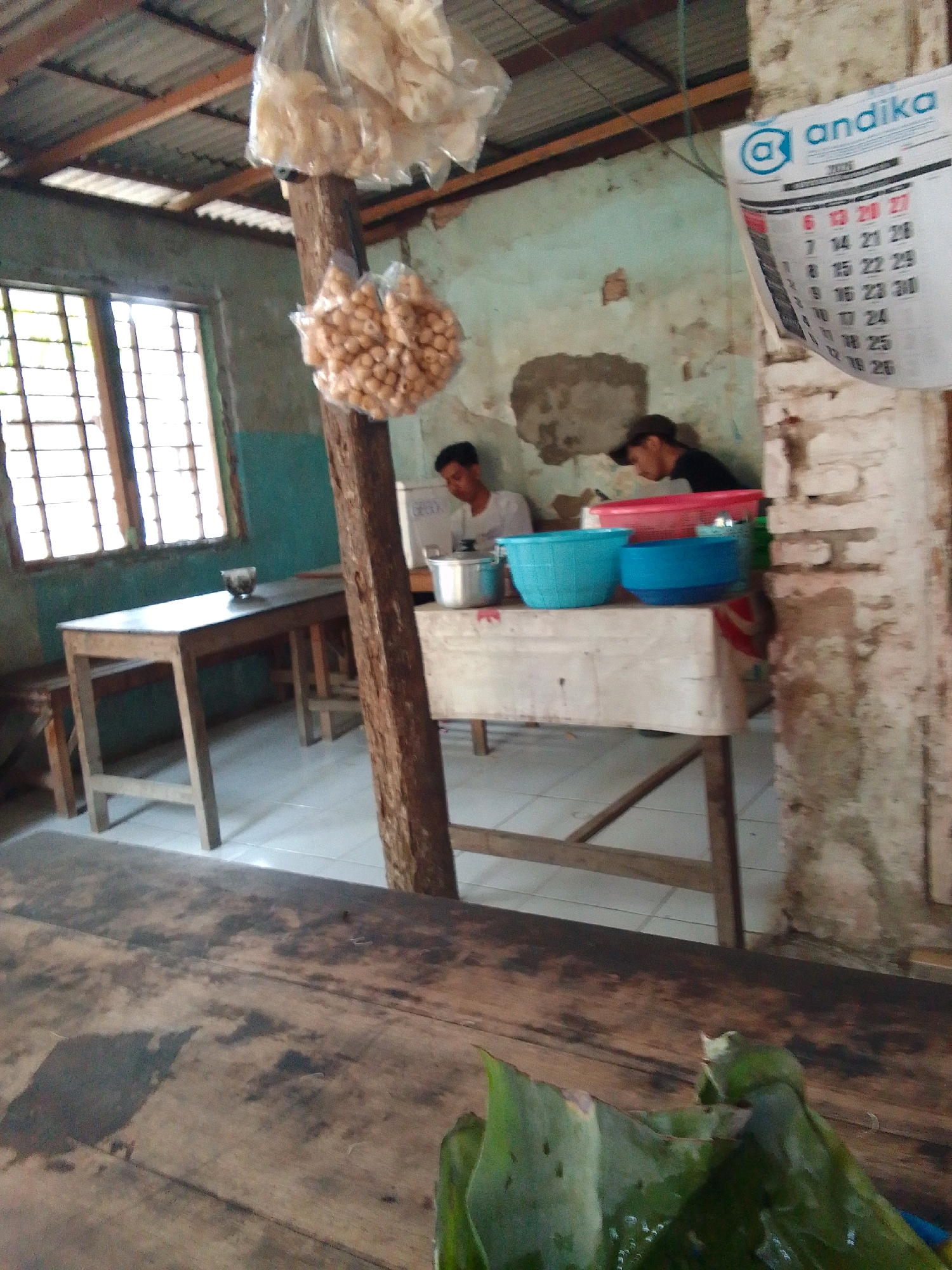 The image depicts a humble, indoor setting, likely a warung or small eatery in Trenggalek, Indonesia. In the foreground, a weathered wooden table with visible stains and grain fills the frame. To the left, a simple wooden table and bench are visible near a window with a grid of panes, suggesting natural light entering the space.
The middle ground reveals a wooden pillar supporting a corrugated metal roof. Hanging from this pillar are two bags of what appear to be snacks or crackers. A larger wooden table is positioned against a wall adorned with peeling paint and textured plaster. On this table, two individuals are present. One, wearing a dark cap and black t-shirt, is seated and leaning forward, potentially engaged in a task or conversation. Another person, wearing a white shirt, is also seated, with their face partially obscured.
On the table in front of the individuals are several items: a silver pot, two blue plastic bowls, and a pink plastic basin. A calendar, displaying the "andika" logo and numbers, hangs on a brick wall in the background, further contextualizing the location. The overall environment appears unpretentious and functional, indicative of a local establishment.
