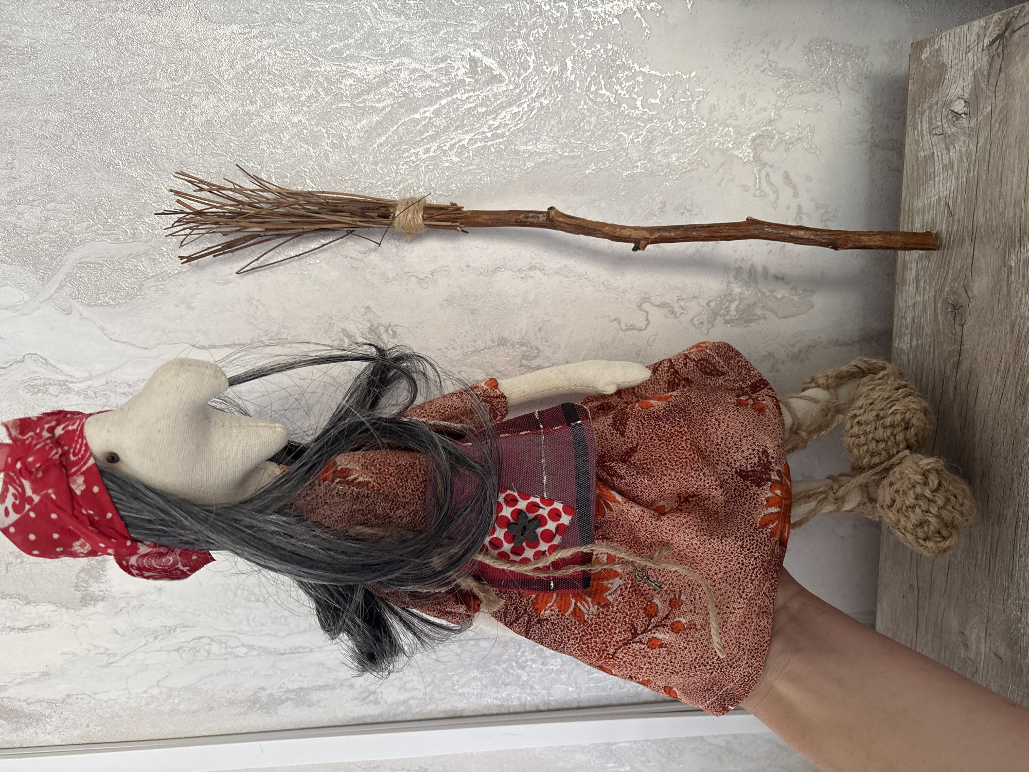 A handmade doll, dressed as a witch with a red bandana and a patterned dress, stands next to a broomstick on a wooden surface against a textured wall. A person's hand is visible holding the doll from the side. The scene is indoors, with no discernible features indicating a specific location in Sochi, Russia.