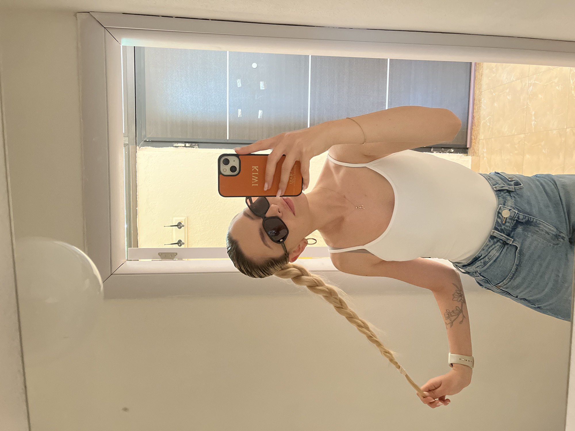 A mirror selfie showing a woman in Unknown, Unknown. She wears sunglasses, a white tank top, and blue jeans. Her blonde hair is styled in a long braid. She is holding an orange smartphone with her name printed on the back in reflective mirror in what appears to be a bathroom.