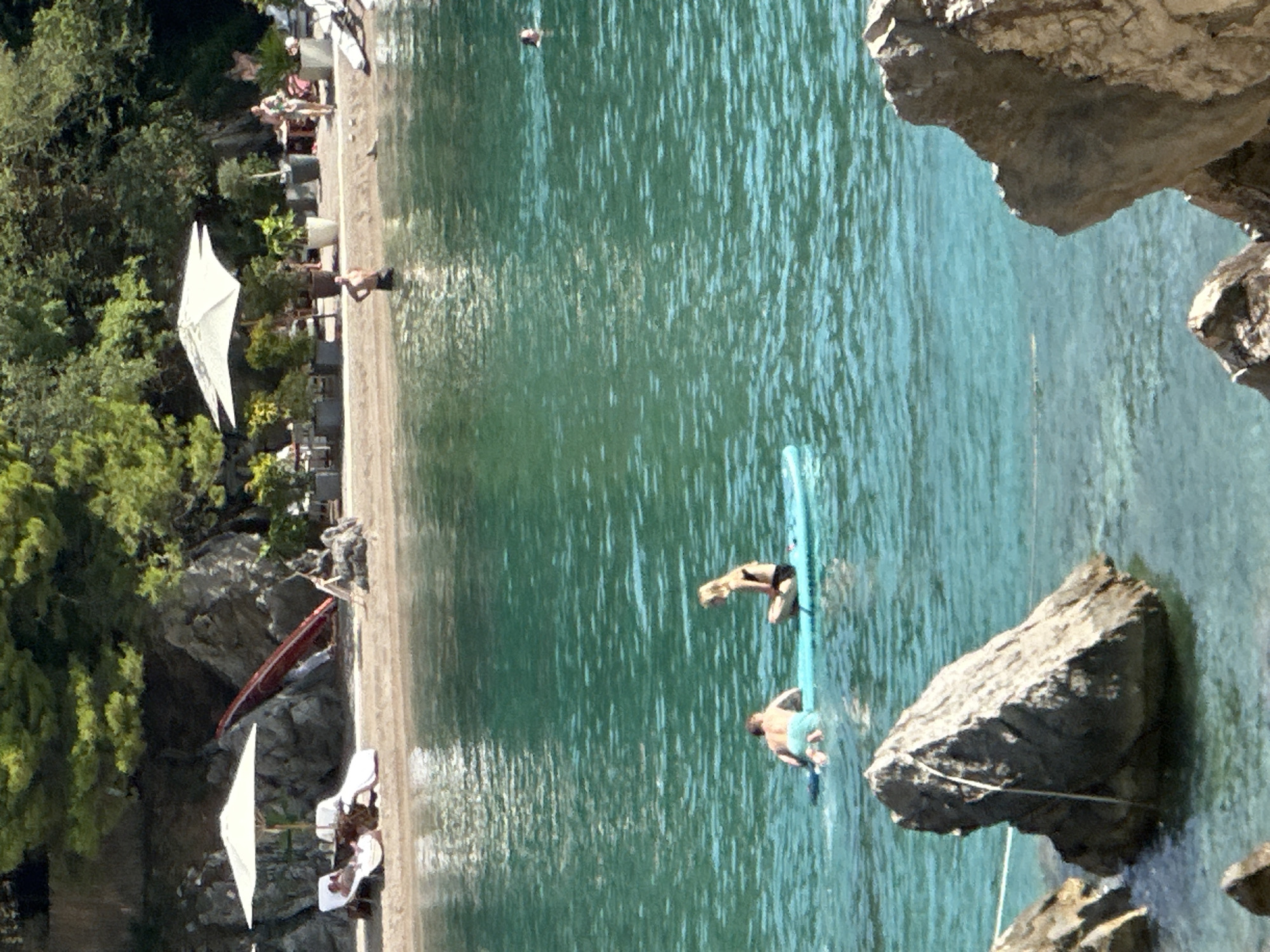 In Budva, Montenegro, a serene beach scene unfolds with clear, turquoise water lapping at a sandy shore. Two individuals are visible in the water: one, a person on a paddleboard, sits calmly, while another person, a male wearing blue swim trunks, paddles nearby. The beach area features lounge chairs and umbrellas, suggesting a recreational space. Further up the shore, vegetation and rocky outcrops frame the scene, hinting at the natural beauty of the Montenegrin coast. In the distance, a few other individuals are present, some wading in the water and others appearing to be on the beach.