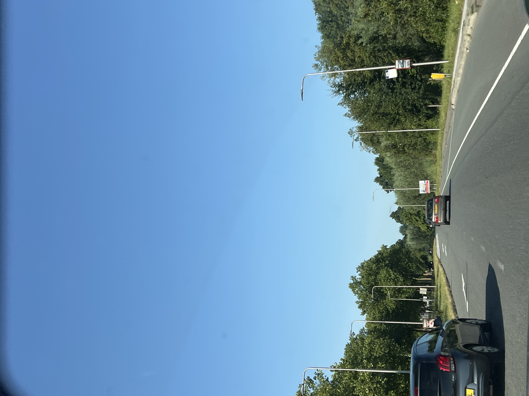 The image depicts a clear, blue sky above a paved road with trees lining both sides. Several cars are visible on the road, moving in the same direction. On the left side of the road, a dark-colored hatchback is in the foreground, with a white directional arrow painted on the asphalt ahead. Further down the road on the left, two streetlights are visible. On the right side of the road, a dark-colored sedan is in motion, and beyond it, more trees and streetlights can be seen. A bus stop sign with multiple panels and a yellow trash bin are situated near the right edge of the road. The scene appears to be a suburban or semi-rural road on a sunny day.