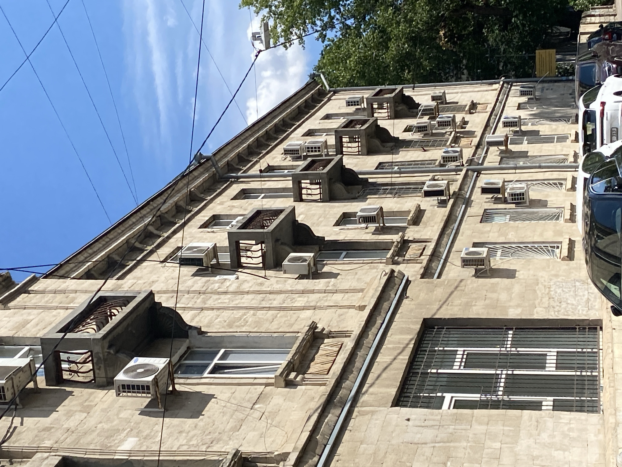 The image shows the facade of a weathered, multi-story apartment building in Chișinău, Moldova, on a sunny day with a bright blue sky. The building's exterior is constructed of light-colored stone blocks, showing signs of age and wear. Numerous air conditioning units are mounted on the walls, attached to windowsills, and on small, ornate balconies with wrought-iron railings. These balconies extend outwards from the building at various levels, some of which are empty, while others hold AC units. The windows are a mix of styles, with some featuring white frames and others with bars. In the foreground, the tops of several parked cars are visible, including a white car with a visible license plate that reads "3JJ". A dense green tree canopy occupies the right side of the frame, and power lines crisscross the sky above the building. The overall impression is of a typical urban residential street scene in an older European city.