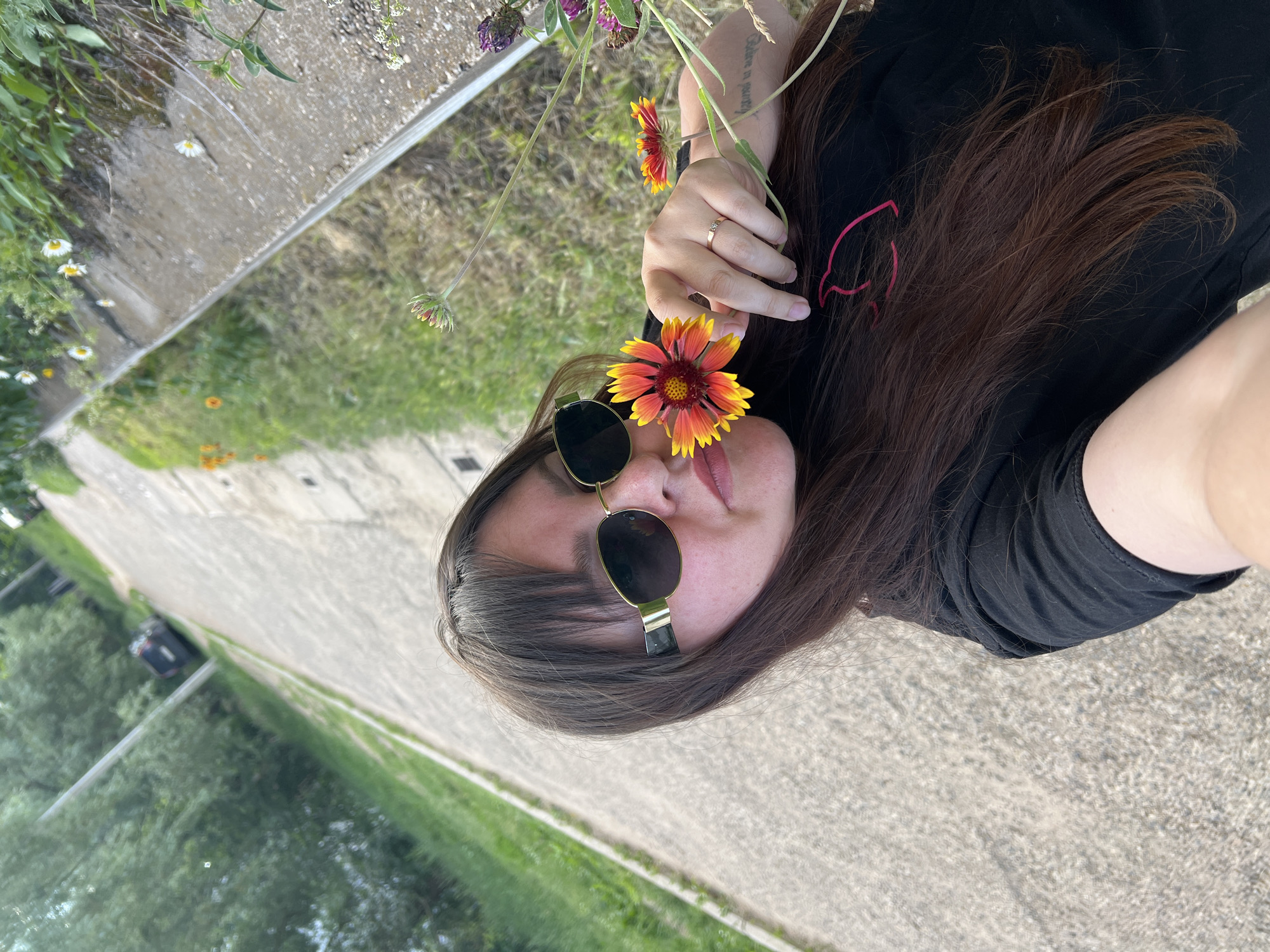 A woman with long, dark hair and sunglasses is taking a selfie outdoors. She is holding a red and yellow flower up to her face, with her right hand visible. She is wearing a black t-shirt. The background shows a paved path and green vegetation, suggesting a park or a roadside. A car is visible in the distance on the path. The lighting indicates it is daytime. The location is identified as Noviy, Russia.