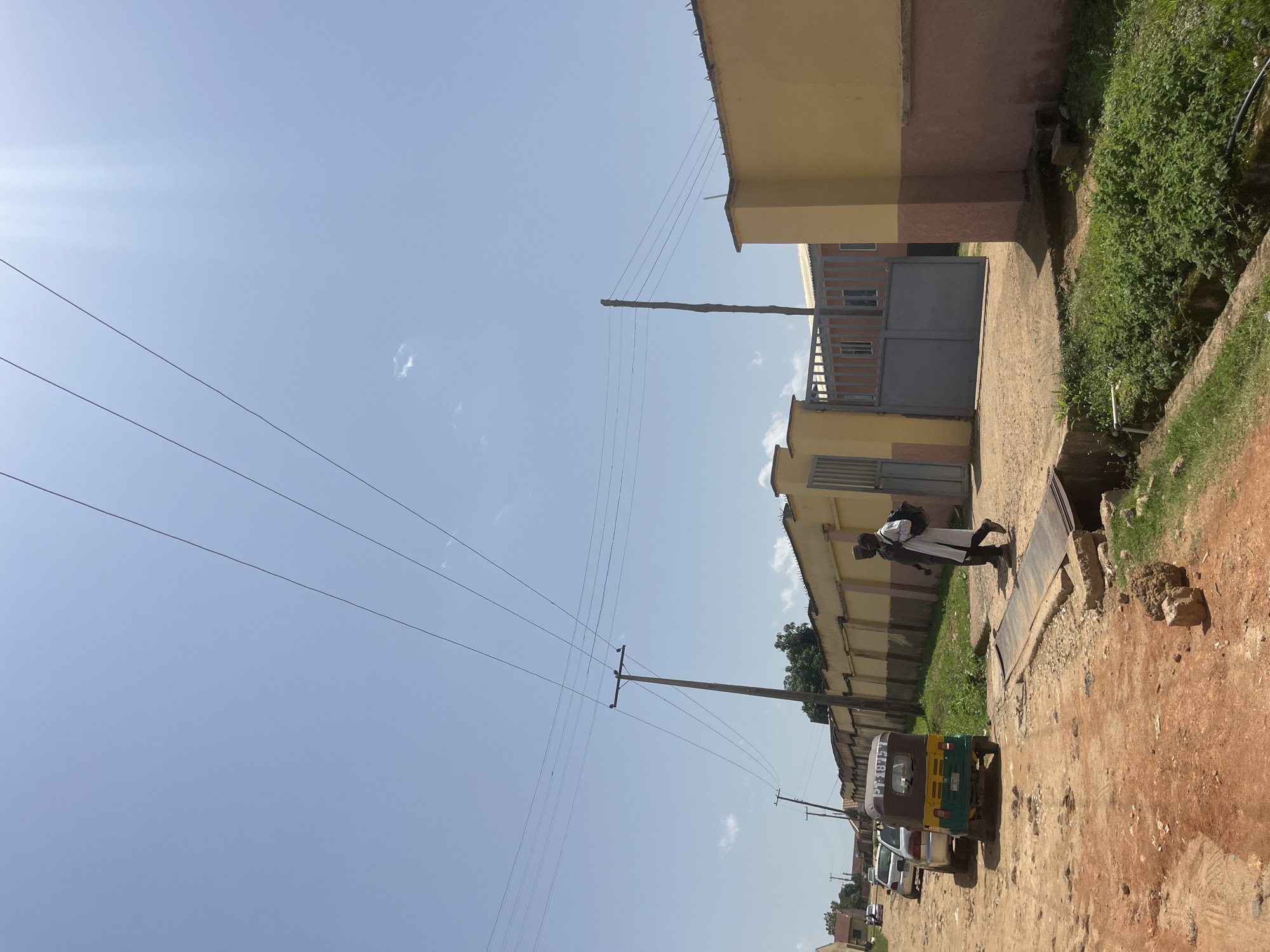 The image depicts a street scene in Gura Top, Nigeria. A dirt road runs through the frame, lined with buildings and a wall. On the left, a colorful auto-rickshaw and two cars are parked. Two individuals, one in a white robe and the other in dark clothing, are walking on the right side of the road. In the background, a yellow building with a brown trim is visible, featuring a large gray gate. Power lines crisscross the clear blue sky, supported by wooden poles. The ground is a mix of red dirt and patches of green vegetation.
