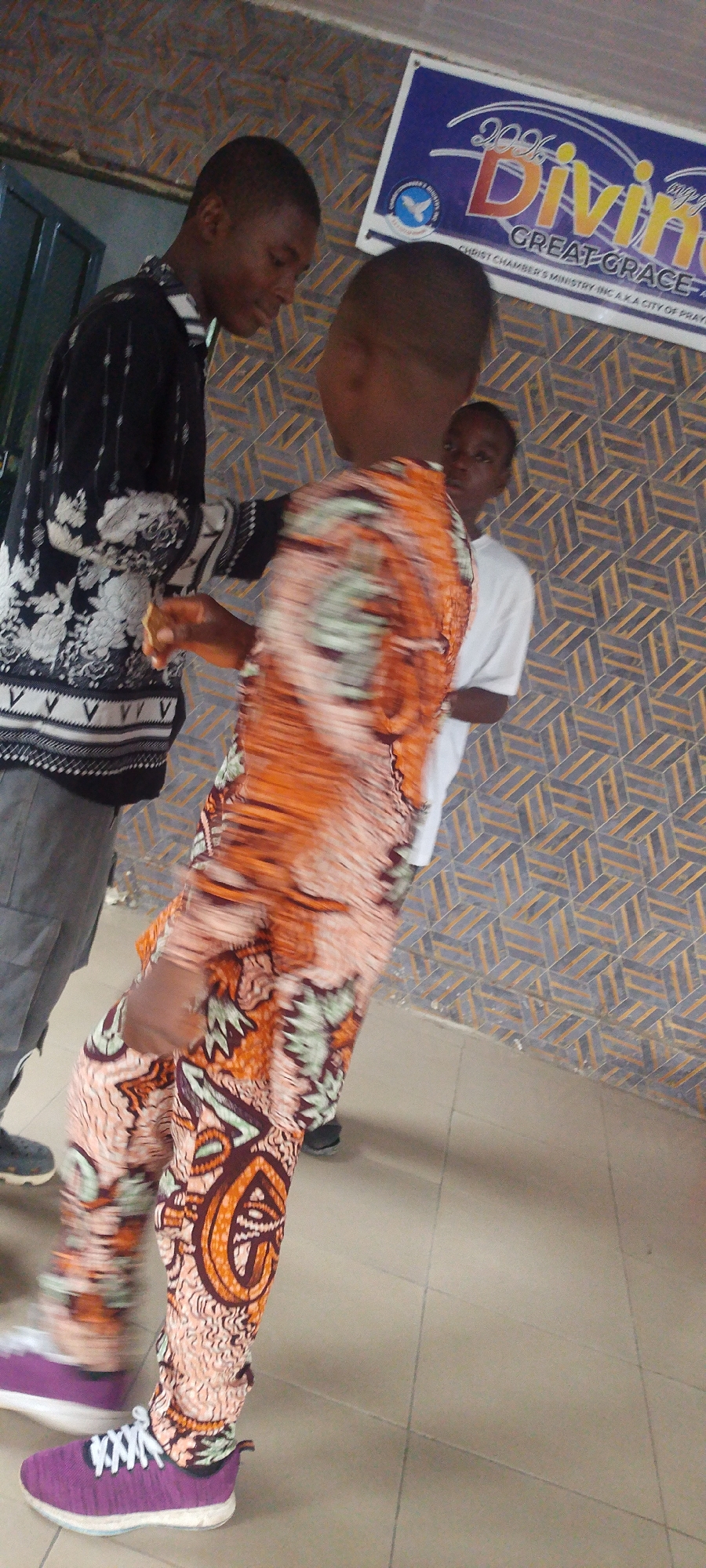 Three young individuals are captured indoors, likely in Lokoja, Nigeria, based on a sign in the upper right corner that reads "CHRIST CHAMBER'S MINISTRY INC A.K.A CITY OF PRAY". The person on the left, appears to be a young man, dressed in a long-sleeved black and white floral patterned shirt paired with grey pants and black shoes. He is looking down, seemingly interacting with the other figure. The person is adorned in a vibrant orange and green patterned outfit consisting of a shirt and pants with purple sneakers. Their features are obscured due to motion blur. Further back, a third person is visible wearing a white t-shirt. The background features a geometric patterned wall.