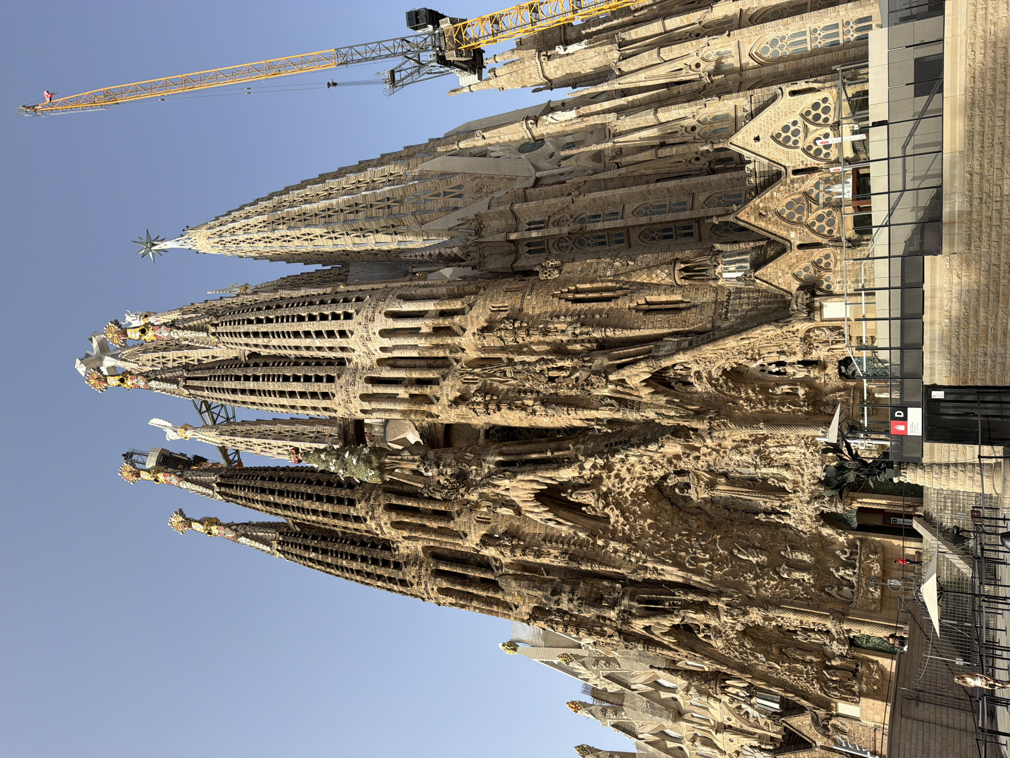 The image shows a low-angle, full-shot view of the Sagrada Familia, a famous Roman Catholic minor basilica located in Barcelona, Spain. The basilica's intricately detailed facade is prominently featured, with its towering spires reaching towards a clear blue sky. A large yellow construction crane is visible to the right of the basilica, indicating ongoing construction or renovation work. Several people are visible in the lower part of the image, including a woman standing near a fence and a person in a red shirt on the steps of the basilica. The environment is sunny, with shadows cast by the building and structures. The overall impression is of a grand and historic architectural site with visible signs of modern development.