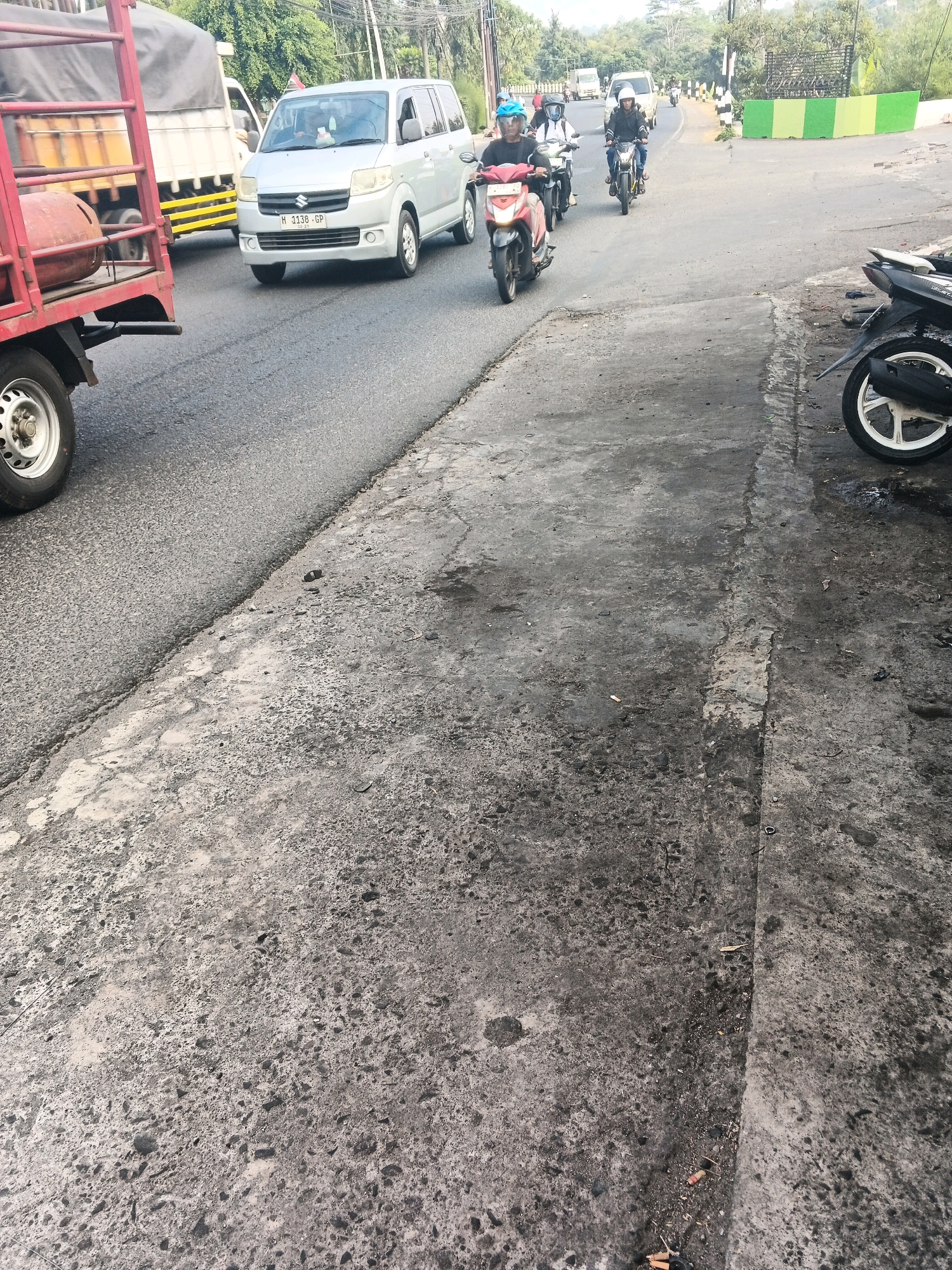 A street scene in Ungaran, Indonesia, shows a traffic intersection. In the foreground, the cracked asphalt road surface is visible, with a parked black and white motorcycle on the right. Further down the road, a silver Suzuki van with license plate H 1138 GP is turning. A red truck, partially visible on the left, is also present. Several individuals on motorcycles are navigating the intersection, with some wearing helmets. The background features trees and buildings, suggesting an urban or suburban environment. The overall lighting indicates it is daytime, possibly late afternoon given the shadows.