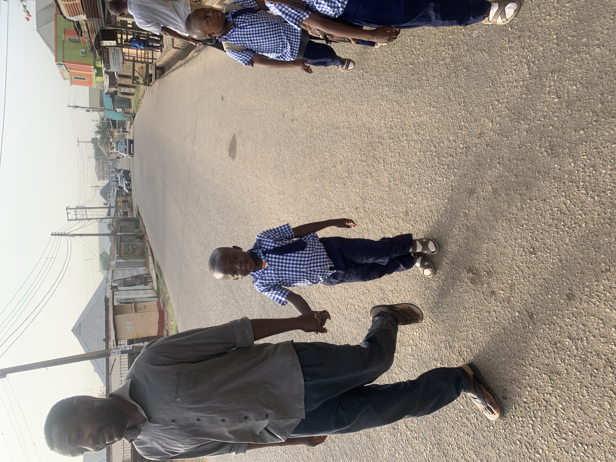 A street scene in Jos, Nigeria, features a man in a grey button-down shirt and dark pants holding hands with a young boy wearing a blue and white checkered uniform and dark pants. They are walking on a paved road. The background includes buildings, utility poles, and vehicles, suggesting a residential or commercial area. There are other children wearing the same uniform ahead of them.