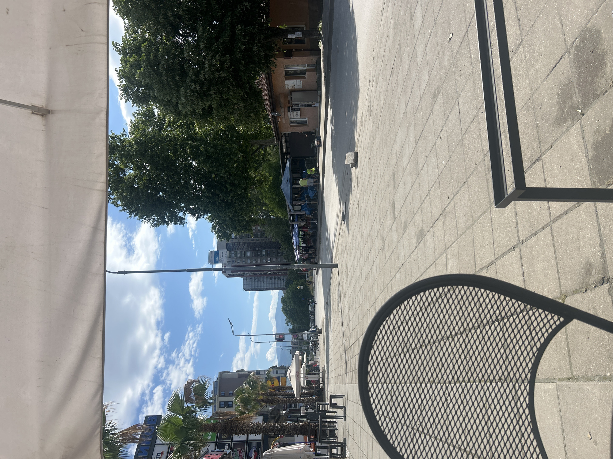 The image depicts an outdoor cafe in Пловдив, Bulgaria, situated on a tiled pedestrian walkway. The view is angled upwards and towards the horizon.
Foreground: Portions of outdoor cafe furniture are visible, including a white canvas umbrella on the left and a patterned metal chair in the lower right corner, casting a shadow on the walkway.
Midground: The pedestrian walkway stretches into the distance. On the left side of the walkway are trees, buildings, and shops.
Background: High-rise buildings are visible in the distance, with clear blue skies and scattered clouds above. Street lights line the walkway.