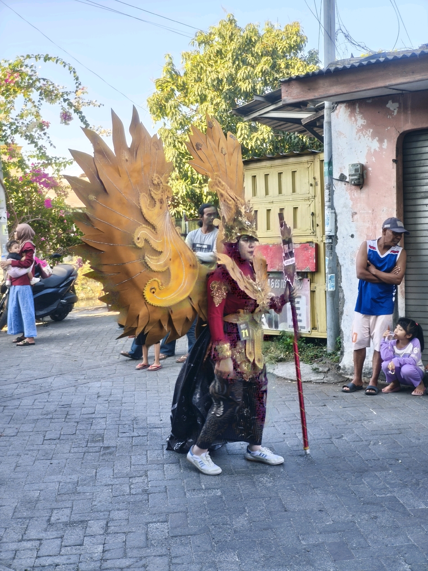 The image depicts a street scene in Kepuh Kiriman, Indonesia, likely during a cultural event or parade. In the foreground, a person in elaborate costume, adorned with large golden wings and a headdress, stands holding a tall staff. The costume features red, gold, and black elements. The person is wearing white athletic shoes.
Several other individuals are present in the background. To the left, a woman in a hijab holds a baby, and a dark-colored motorcycle is parked nearby. Further back, a man in a grey t-shirt is visible. To the right, a man in a blue sleeveless shirt and a cap stands with his arms crossed, looking towards the costumed individual. A young girl in purple clothing sits on the ground beside him, looking up.
The environment consists of a paved street with a brick-like pattern. Buildings with corrugated metal roofs and weathered walls line the street. A yellow gate is also visible. Lush green trees and flowering plants contribute to the outdoor setting. The sky is clear and blue, suggesting a sunny day. The overall atmosphere appears to be one of community gathering and celebration.