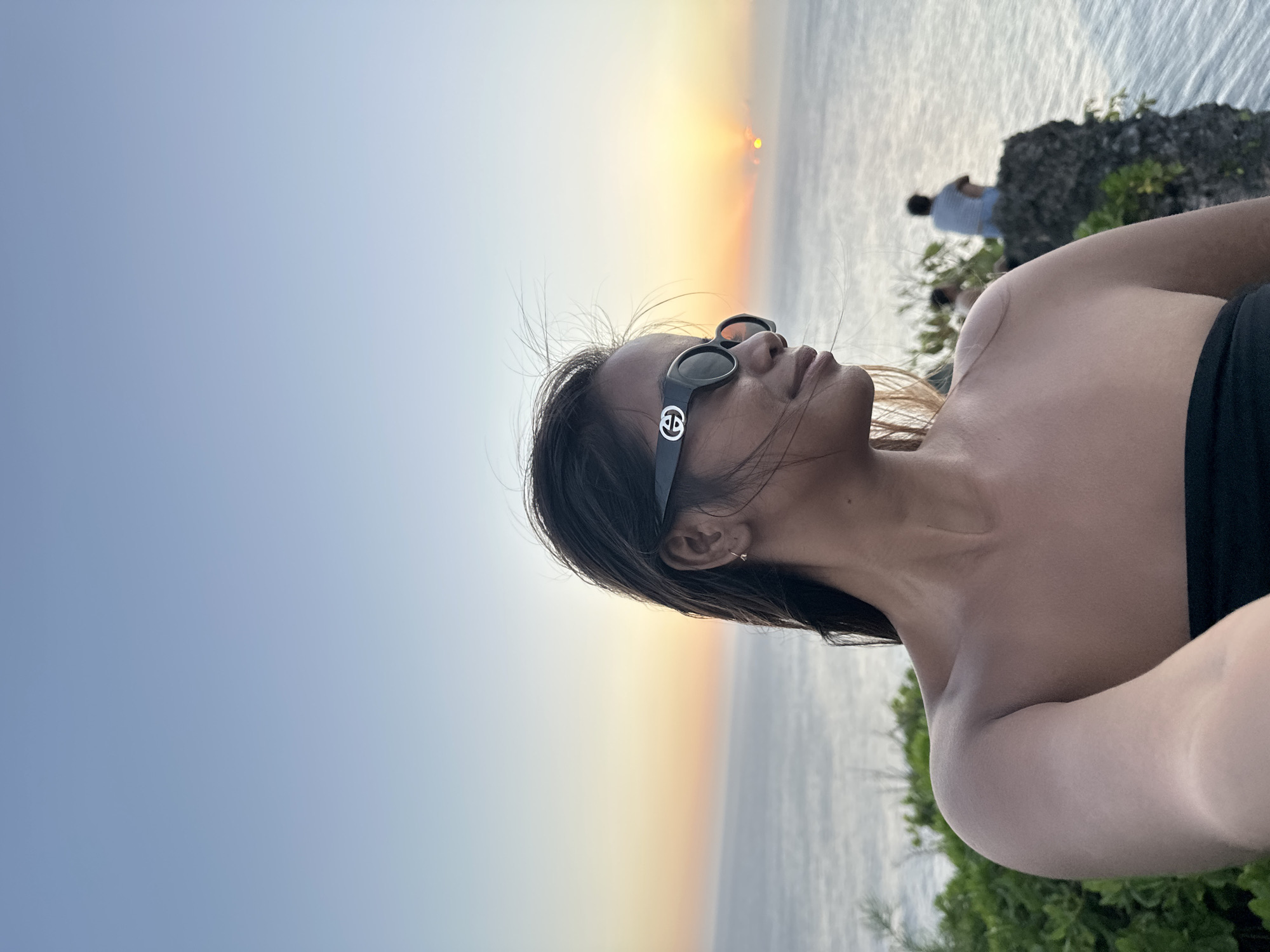 An eye-level medium selfie shows a woman with dark hair wearing black sunglasses with a Gucci logo on the right side of her face, looking to her right. She is wearing a black strapless top, and her bare shoulders are visible. The background shows a body of water, likely the Indian Ocean, with the sun setting on the horizon. Two silhouetted figures are sitting on a rock formation to the right, in the distance. The scene is set on a cliff overlooking the ocean in Kuta Selatan, Indonesia, characterized by lush green vegetation in the foreground.