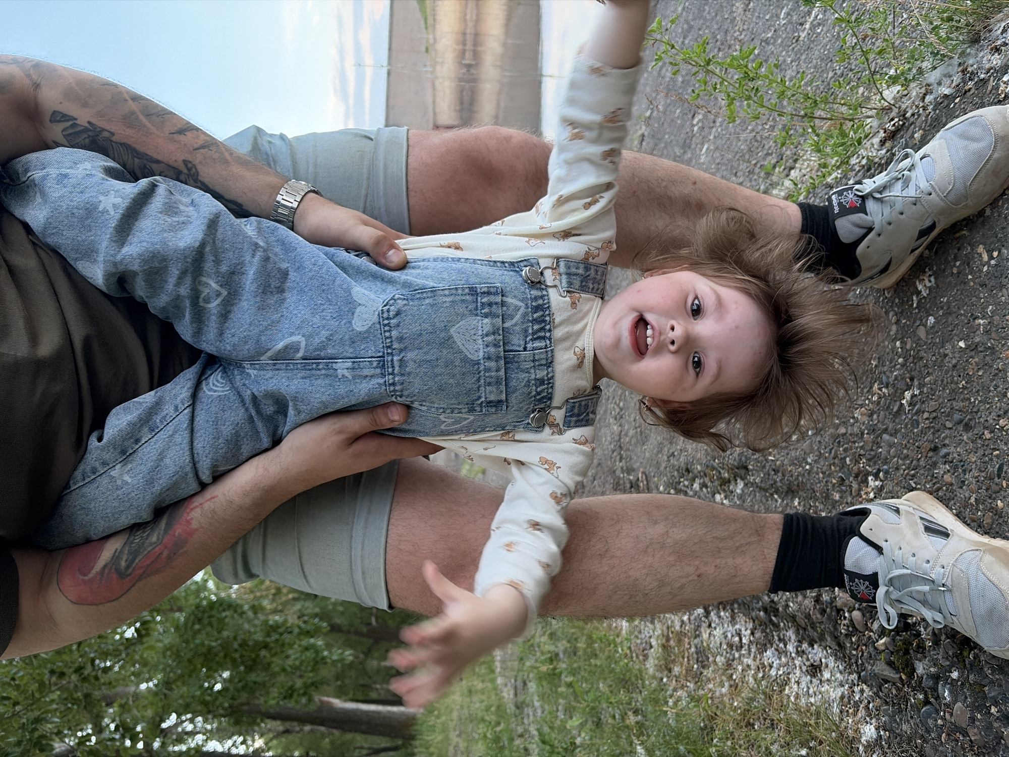 In this outdoor, eye-level, medium shot, a person is holding a young child upside down by the legs. The child, wearing a light-colored shirt with a teddy bear pattern and denim overalls, has a smiling expression and their arms are extended. The adult is wearing a grey t-shirt, grey shorts, black socks, and light-colored athletic shoes. Tattoos are visible on the adult's arms. The background shows a gravel path, green trees, and a body of water with a dam structure. The scene appears to be located at the Kama River embankment in the city of Chaikovsky, Russia.