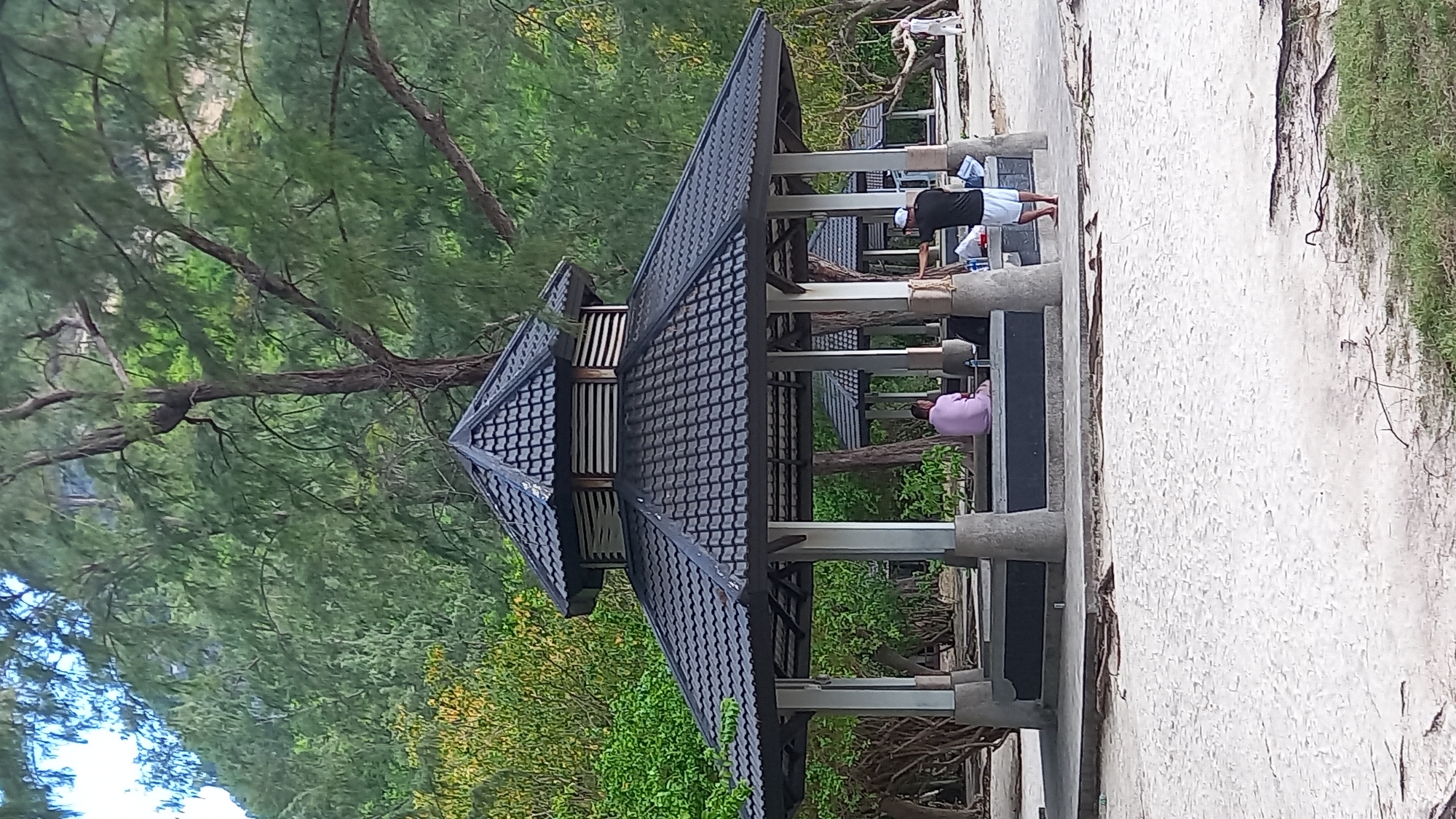 The image shows a gazebo located on a beach in Ban Ao Nang, Thailand. The gazebo has a dark tiled roof and is supported by concrete pillars. A person wearing a pink shirt is sitting inside the gazebo. Another person wearing a black shirt and white shorts is standing on the edge of the gazebo, appearing to be stretching or doing exercises. The surrounding environment includes trees, greenery, and the sandy beach.