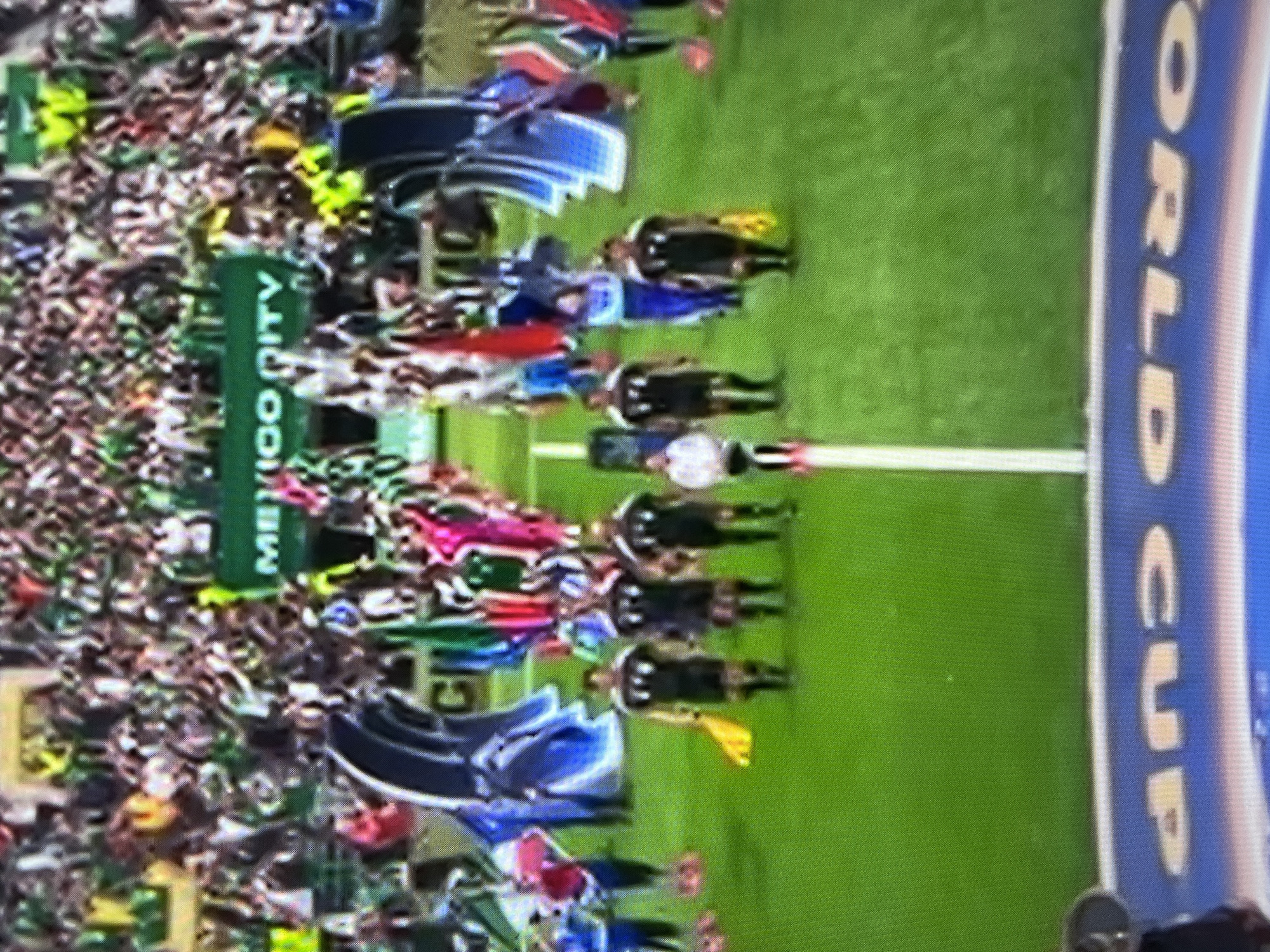 A group of individuals, some wearing black uniforms with white trim and others in civilian attire, walk across a green field. Two individuals in the foreground, dressed in black uniforms, are holding yellow flags. Behind them, a larger crowd of spectators, predominantly dressed in green, occupies tiered seating. A green banner with white text reading "MEXICO DITY" is visible in the background. In the lower portion of the image, a blue surface with white text that appears to read "WORLD CUP" is present. The scene suggests a sporting event, possibly a football (soccer) match, with officials and participants on the field and a crowd in the stands.