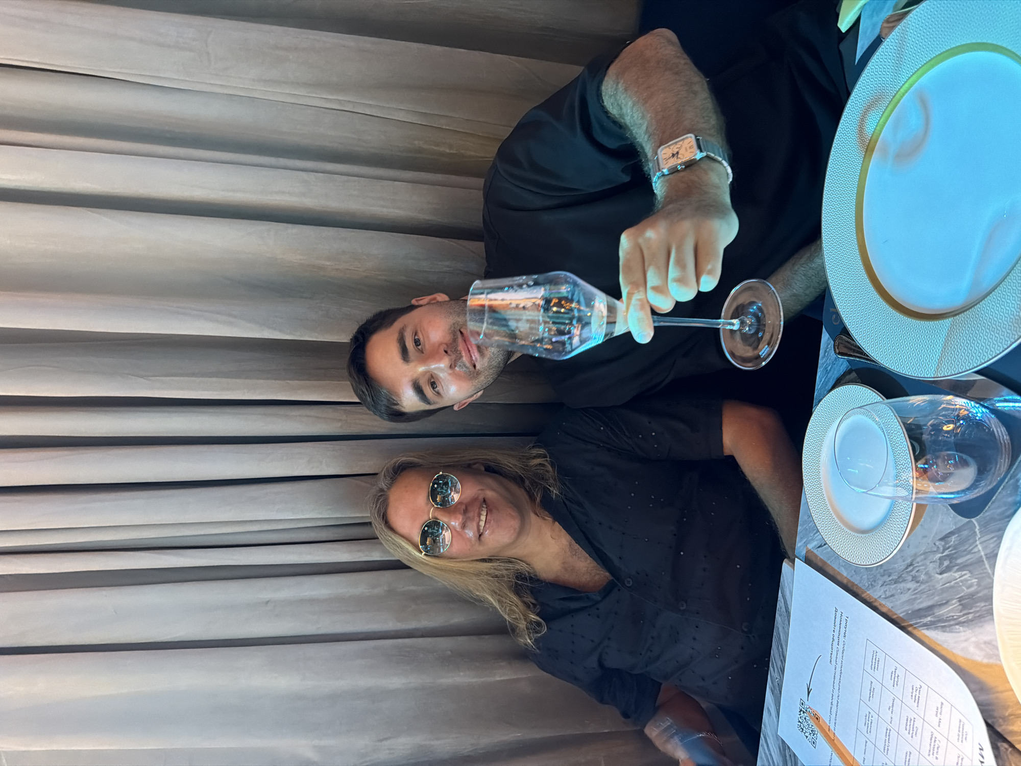 Two men are seated at a table in an indoor setting. The man at the top of the image is holding a champagne flute, wearing a watch, and dressed in black. The man at the bottom of the image has long blond hair and is wearing sunglasses and a black button-down shirt. The table is set with plates and a menu or document with a QR code is visible. The background features a curtained wall. This scene is located in Choeng Thale, Thailand.