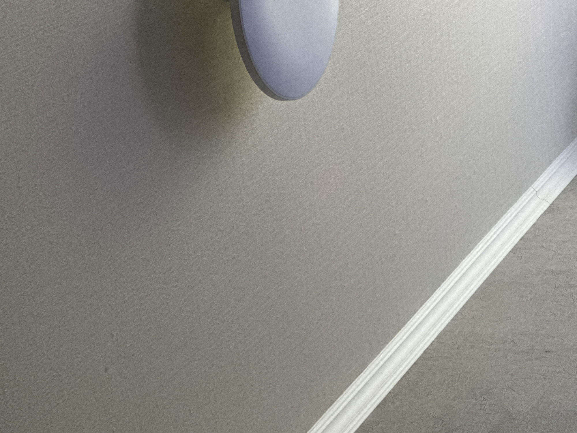 The image shows a close-up of a textured gray wall with a white decorative molding strip running diagonally from the bottom right corner towards the top. The molding has a multi-layered, carved design. In the upper left portion of the image, a portion of a white, oval-shaped object is visible, casting a shadow on the wall. This object appears to be a light fixture or a similar wall-mounted item. The lighting suggests it is daytime, possibly indoors, given the even illumination of the wall and the distinct shadow. The texture of the wall is visible, appearing to be a wallpaper with a subtle pattern. No people, text, or specific environmental cues like weather are discernible. The setting is a wall interior, likely in a room within a residential or public building. The overall impression is one of a simple architectural detail within an indoor environment.