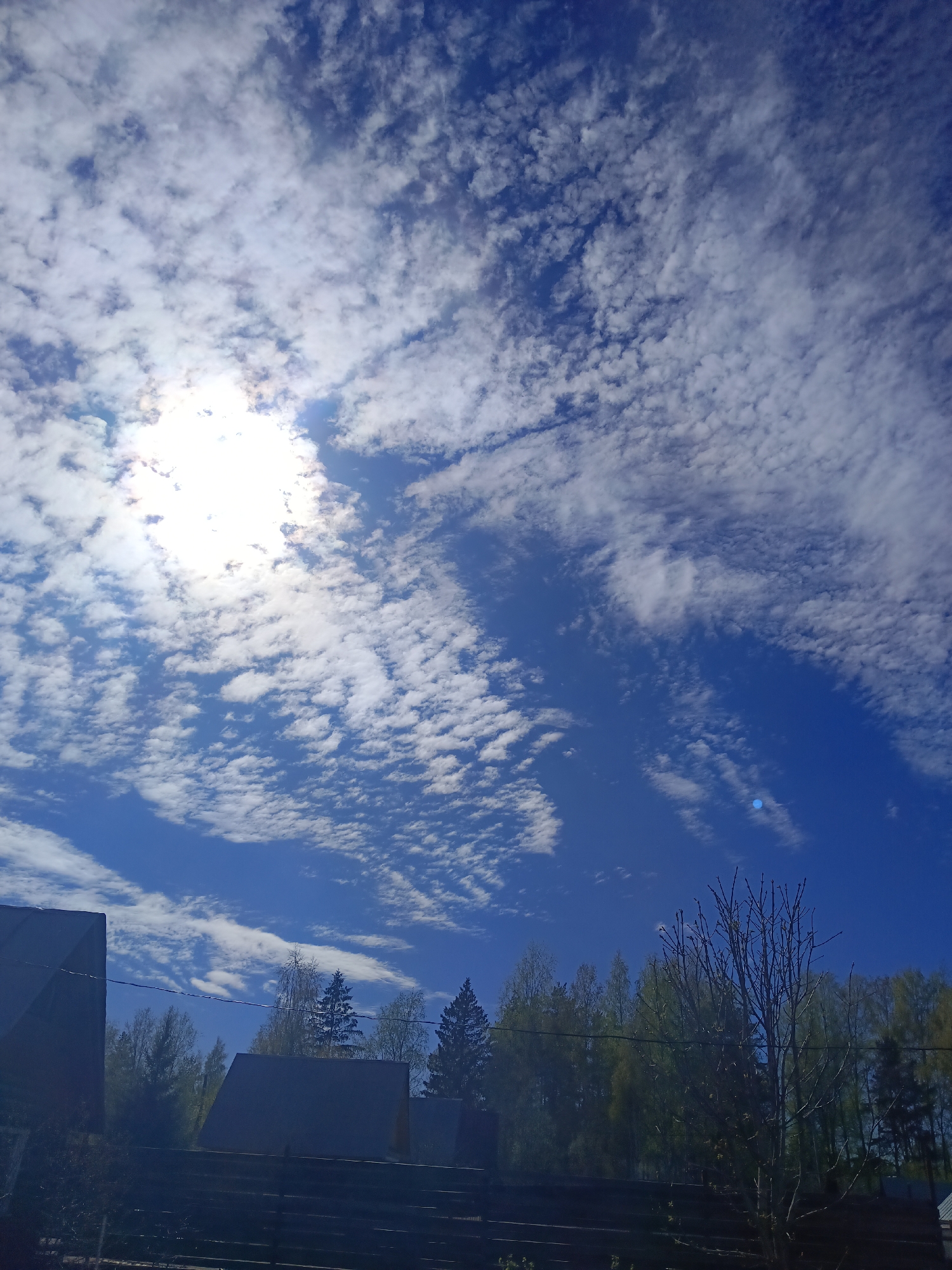 The image depicts a bright, daytime sky with scattered clouds and the sun partially obscured by them. Below the sky, a treeline is visible with a mix of deciduous and coniferous trees. In the foreground, there are rooftops of buildings and a fence. The location is Вичелово, Russia.