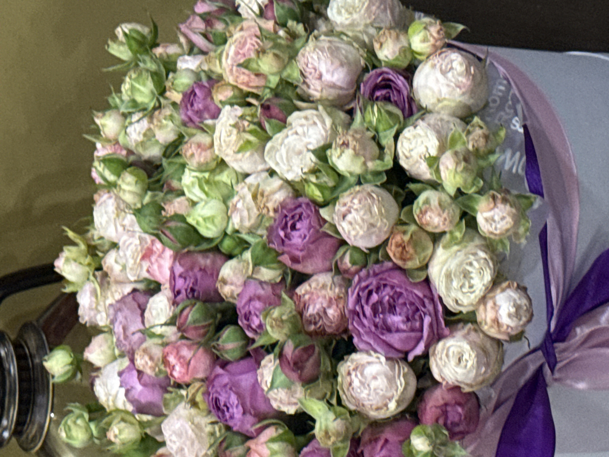 The image depicts a bouquet of roses, mostly in shades of white and purple, with some green buds visible. The bouquet is tied with a ribbon in coordinating shades of purple. The setting appears to be indoors, and the background is indistinct. Although the image shows a bouquet of roses, there is no evidence to confirm that it is located in the city of Yerevan, Armenia.