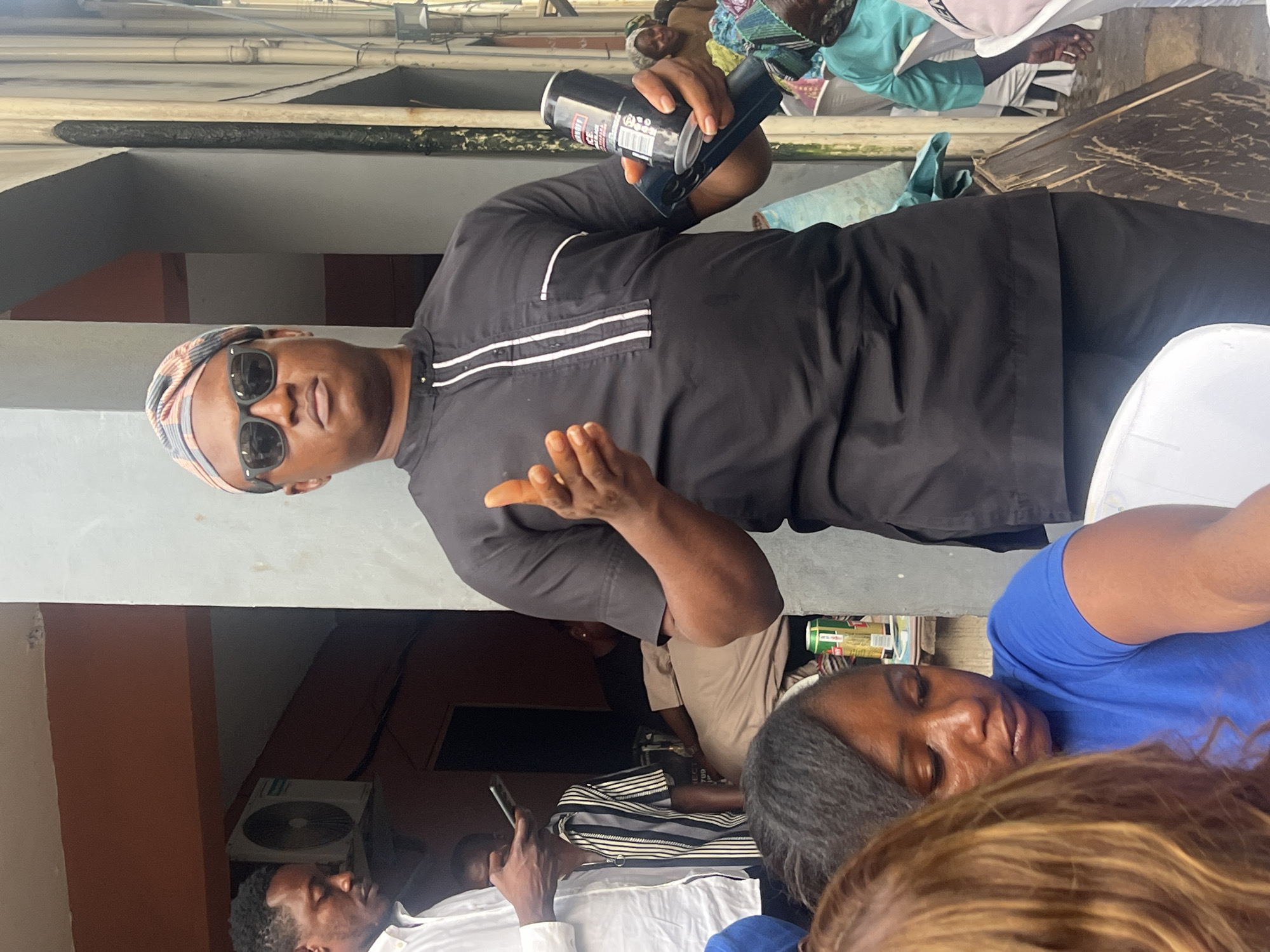 Here's a detailed description of the scene:
**Location:** The photograph was likely taken outdoors in Lagos, Nigeria, as suggested by the style of architecture and attire of the people.
**People:** A man in sunglasses and a black shirt with white stripes on the shoulders is holding up a black can. He also has a patterned head covering. There are other people in the image, including a woman in a blue shirt, and other people in the background.
**Objects:** The man is holding a can with a black label. Other objects include white chairs, cans of drinks, cell phones, and some electrical equipment outside a building.
**Actions:** The man is gesturing with his free hand, and holding the can, possibly presenting or describing it to the group.
**Interactions:** The man with the can appears to be addressing a group of people.
**Environment:** The setting appears to be an outdoor or semi-outdoor space, possibly a patio or courtyard next to a building. There is some vegetation visible and a building in the background with an air conditioning unit.