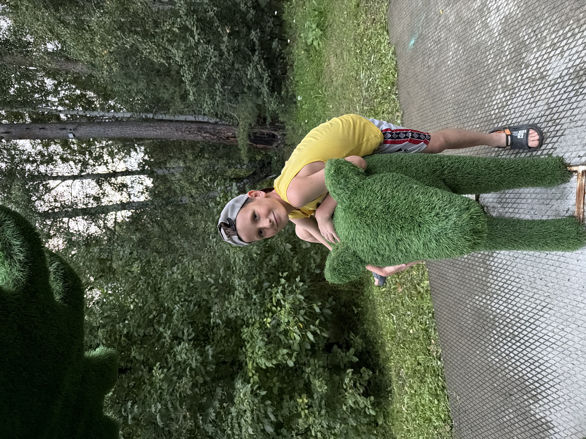A young boy wearing a yellow tank top, gray and white baseball cap, and patterned shorts leans on a large, green, artificial grass sculpture of an animal, possibly a dog or a deer, with its ears up. He is outdoors, in what appears to be a park or recreational area. The background shows lush green trees and foliage, with tall, slender tree trunks visible. The ground in the foreground is a grey, metal grate, likely part of a pathway or play structure. The lighting suggests it is daytime, possibly late afternoon or early evening. This scene could be located in the city of Zarechny, Russia, given its appearance as a green, public outdoor space.