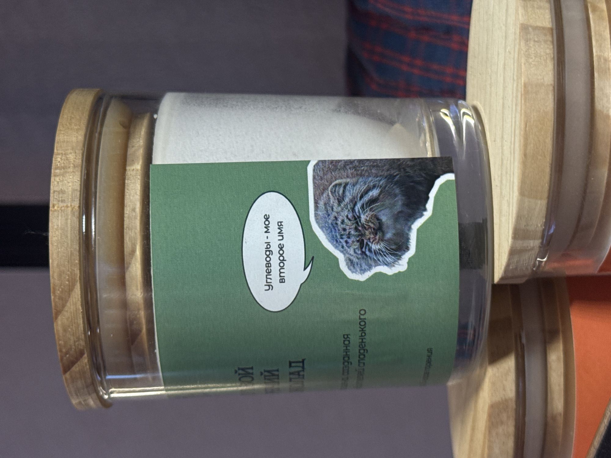 The image shows a cylindrical glass jar filled with a white substance. The jar is sealed with a wooden lid and has a green label affixed to its side. A cutout image of a cat's face is prominent on the label, along with a speech bubble containing text in Russian: "Углеводы - Мое второе имя" (Carbohydrates - My second name). The jar appears to be placed in front of other similar jars with wooden lids. While the image itself doesn't provide explicit information about the location, the context (given the cat face) suggests that this is from a product being sold at the Izhevsk Zoo in Izhevsk, Russia.