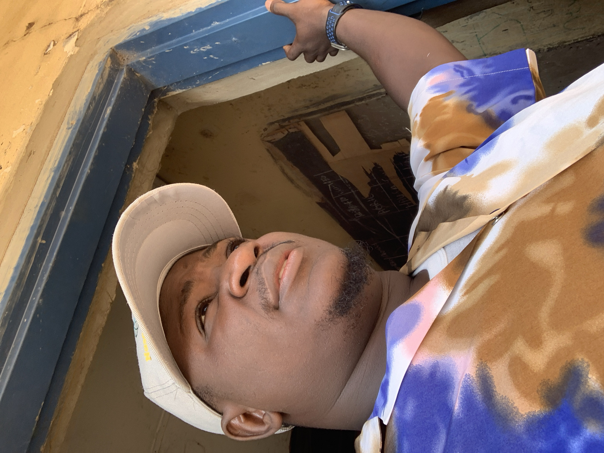 The image shows a man wearing a light-colored baseball cap and a colorful, tie-dye shirt. He is standing in a doorway. His right hand is visible, resting on the doorframe. The doorway appears to be made of blue painted wood and the wall next to it is a light yellow color. In the background, there is a dark surface with some writing on it, possibly a chalkboard. The lighting suggests it is daytime, with light coming from the right and illuminating the man's face from the side. The scene is likely outdoors or in a partially enclosed space, given the light and the rustic appearance of the doorway and wall. The location in Akwanga, Nigeria is suggested by the context provided.